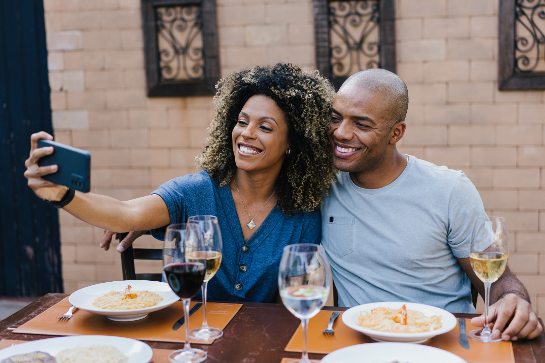 A man and woman sitting at a table outdoors, taking a selfie together, with three glasses of wine and plates of spaghetti in front of them.