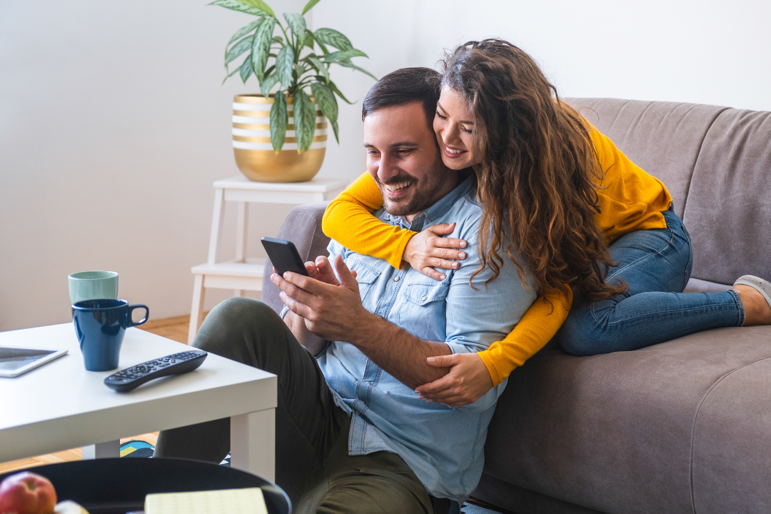 A couple sitting on a gray couch, smiling and looking at a smartphone. The woman is hugging the man from behind. A white table in front of them has two mugs, a remote, and a notebook. There is a potted plant on a small white table in the background.