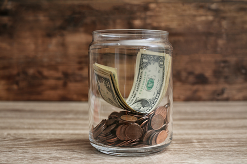 A glass jar filled with dollar bills and coins on a wooden surface.