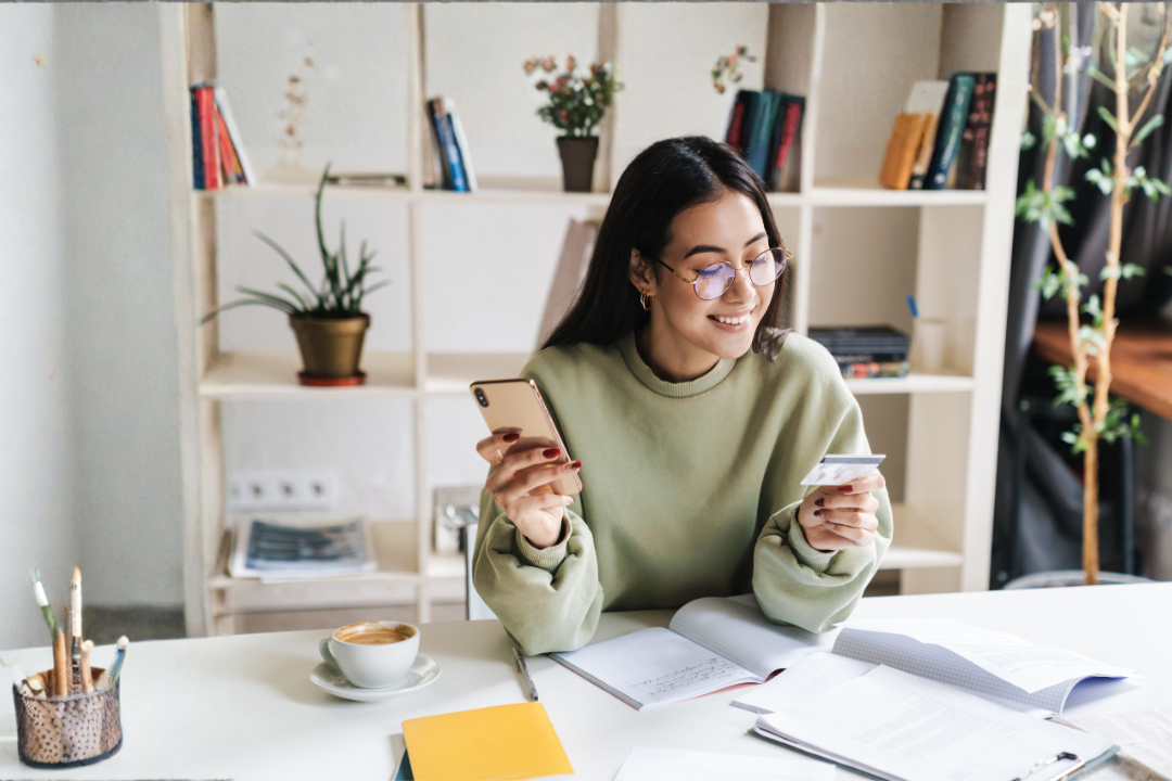 Young smiling woman sitting at her home office desk, holding her cell phone and looking at her debit card.