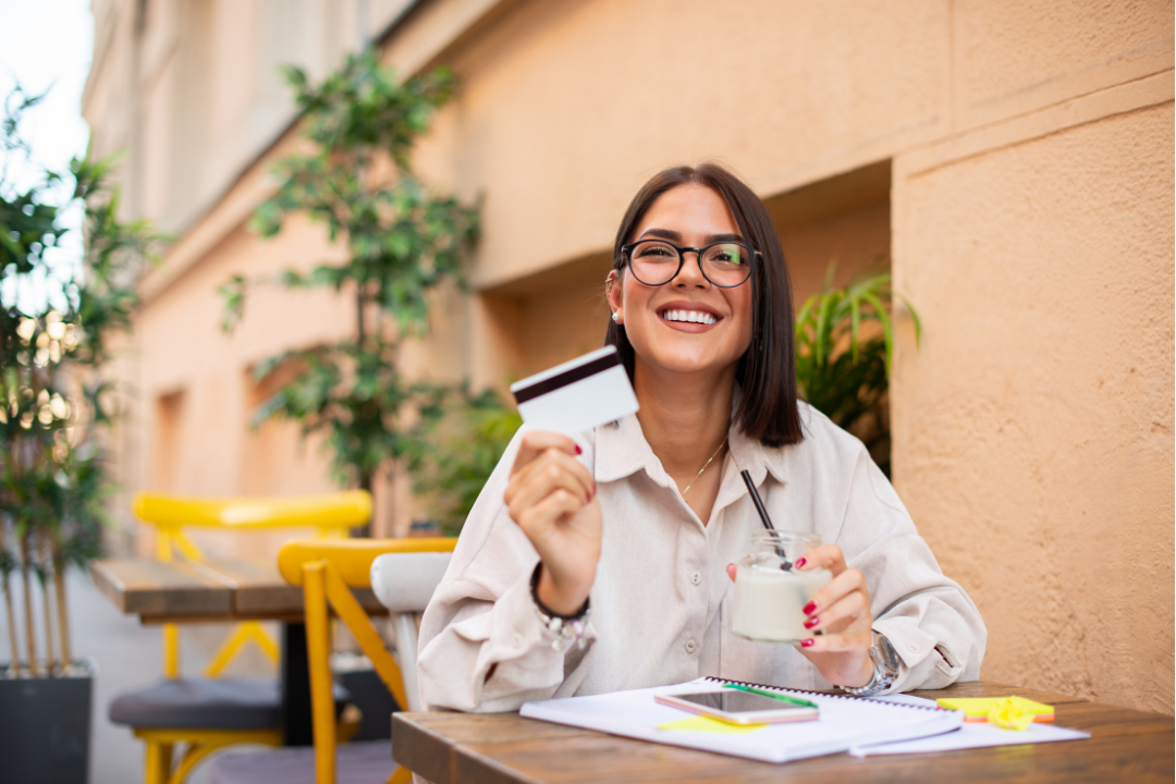 A smiling woman in glasses sitting at an outdoor cafe table, holding a credit card and a jar with a drink, with notebooks, a phone, and highlighters on the table.