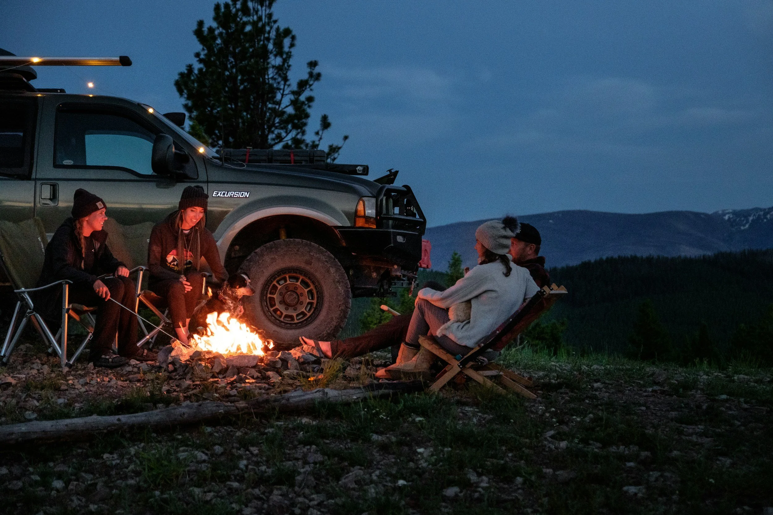 Group of five people sitting around a campfire near a vehicle on a mountain landscape at dusk.