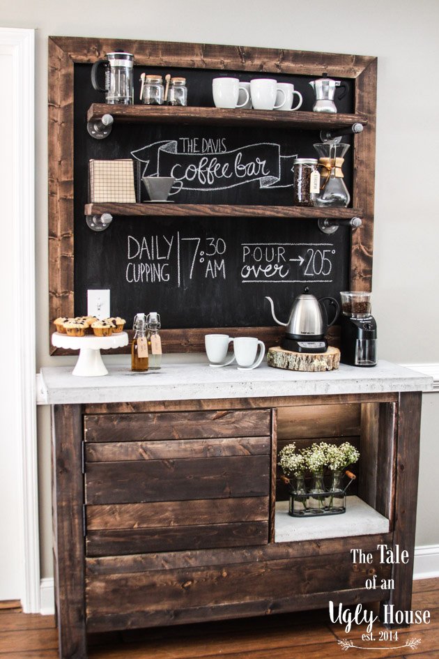 A coffee station with a dark wood and white marble counter, a blackboard with coffee instructions, coffee-making equipment, mugs, cupcakes, and small bottles, against a beige wall.