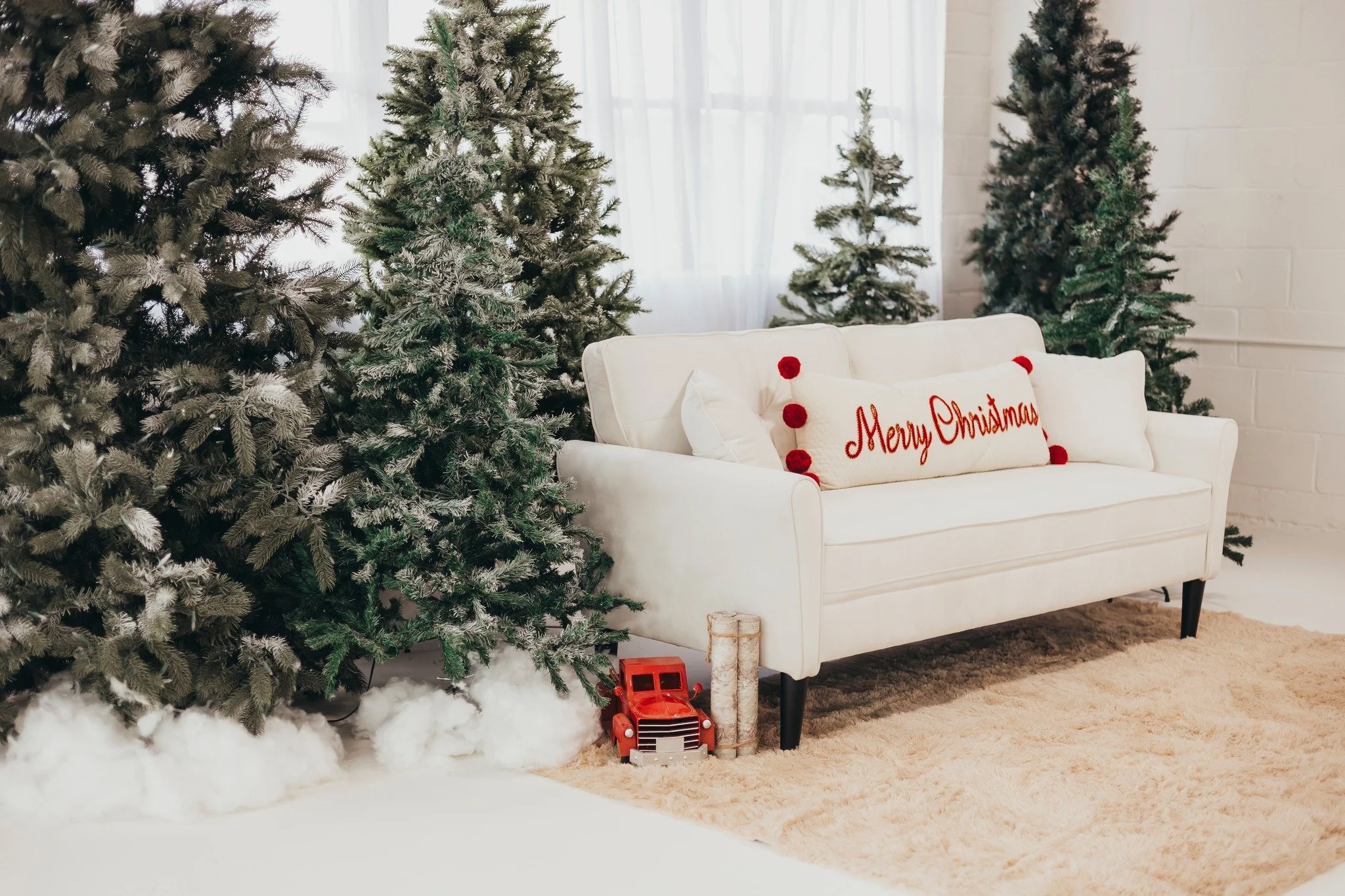 Festive Christmas living room with multiple decorated pine trees, a white sofa with red and white pillows, including one with 'Merry Christmas' embroidered, a red toy truck, and faux snow on the floor.