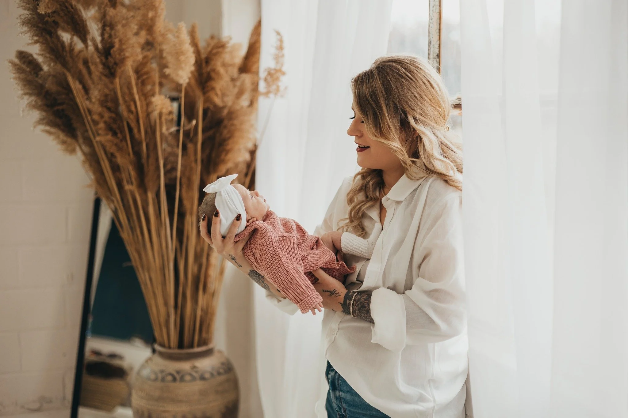 A woman holding a baby girl with a white bow on her head, smiling at her in a bright room with white curtains and a large vase of dried pampas grass.