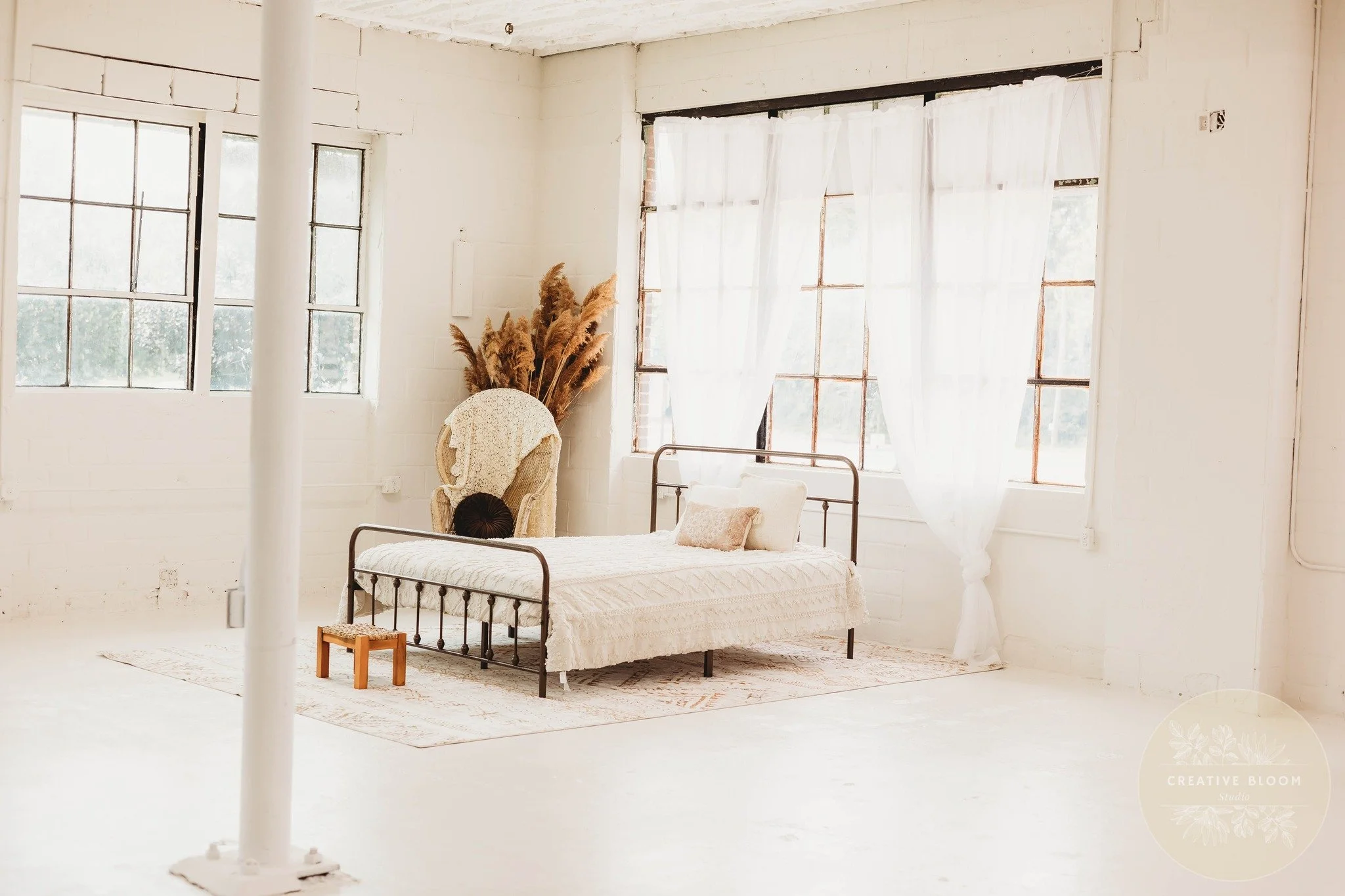 Bright, airy bedroom with white brick walls, large windows with sheer white curtains, a black metal bed with white bedding and pillows, a vintage armchair with a lace cover, a bundle of dried pampas grass next to the armchair, and a small wooden stool.