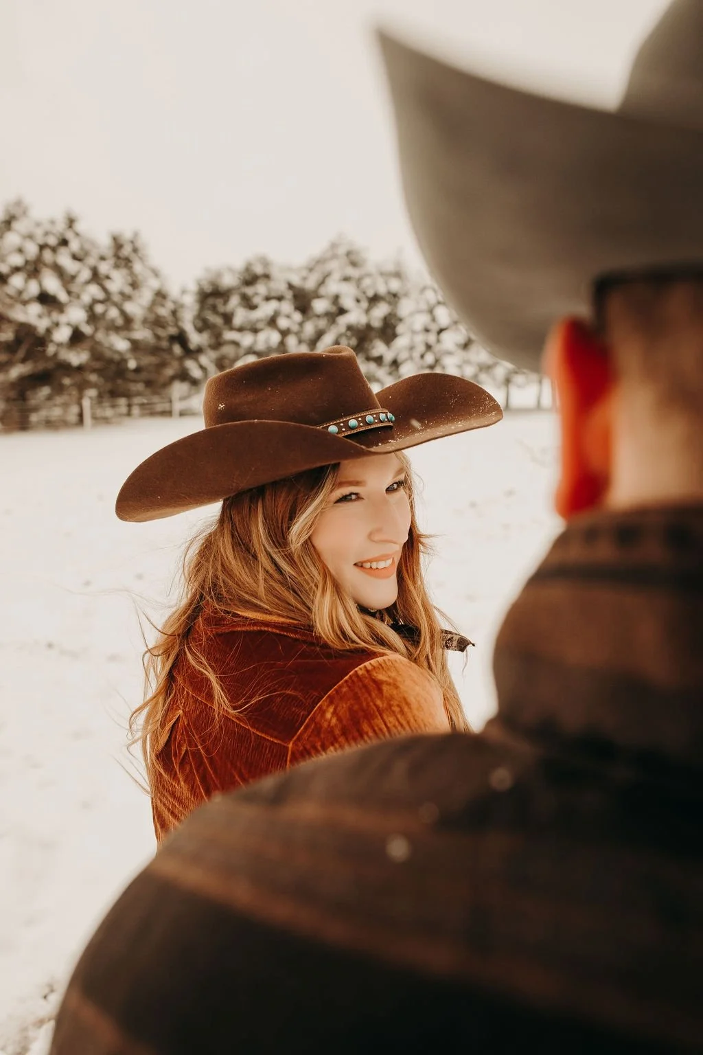 A woman with wavy hair smiling and squinting in a snowy outdoor setting, wearing a brown wide-brimmed cowboy hat with turquoise accents and a brown corduroy jacket, with a person wearing a gray cowboy hat in the foreground.