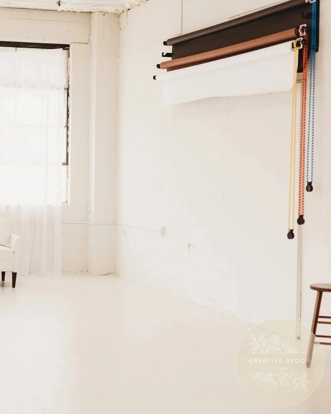 Photo of a minimalist photography studio with a white wall, a large window with white curtains, a white chair, and a backdrop system with brown, black, and white rolls of paper, and measuring tape hanging on the wall.