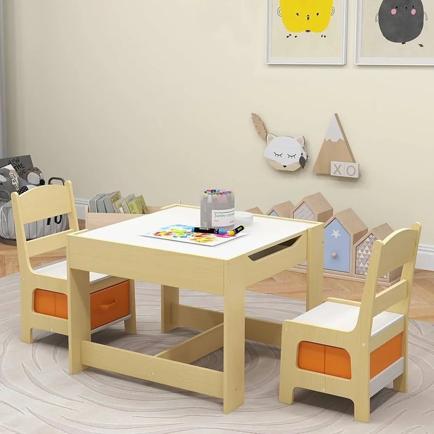 A children's playroom with a small yellow table and two matching yellow chairs with orange accents. The table has coloring supplies on top. The wall behind features framed animal artwork, soft plush wall-mounted decorations including a fox and a mountain with labeled X and O, and a row of small house-shaped shelves in pastel blue and beige. There is a white circular rug on the wooden floor.