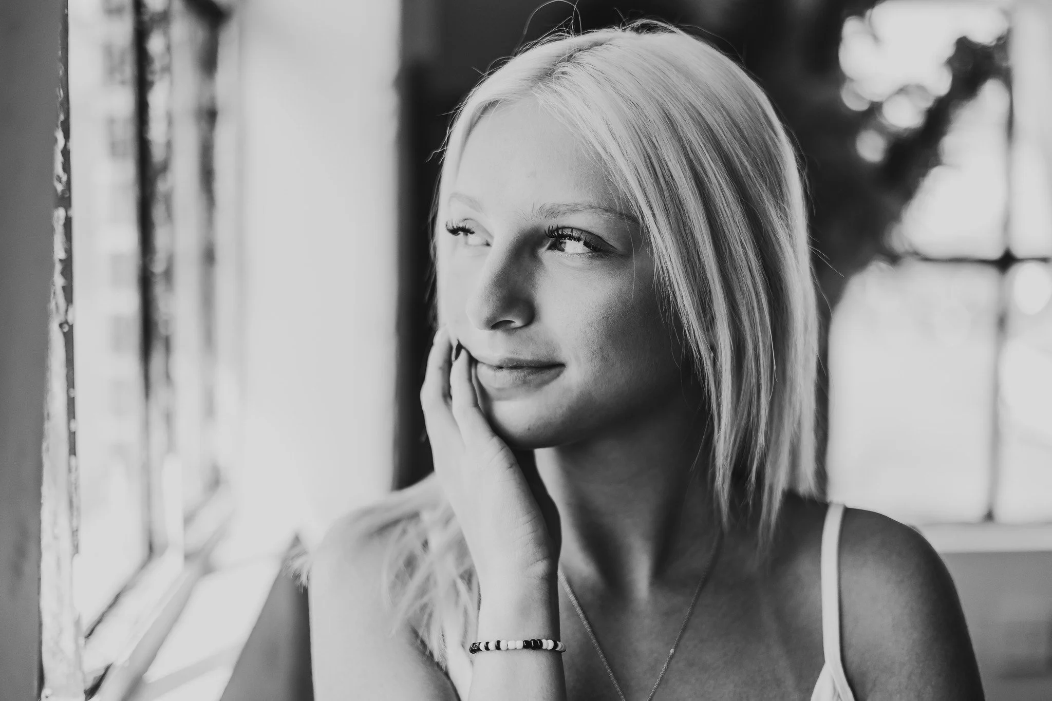 Black and white photo of a woman with blonde hair looking out of a window, resting her face on her hand, with soft light illuminating her face.
