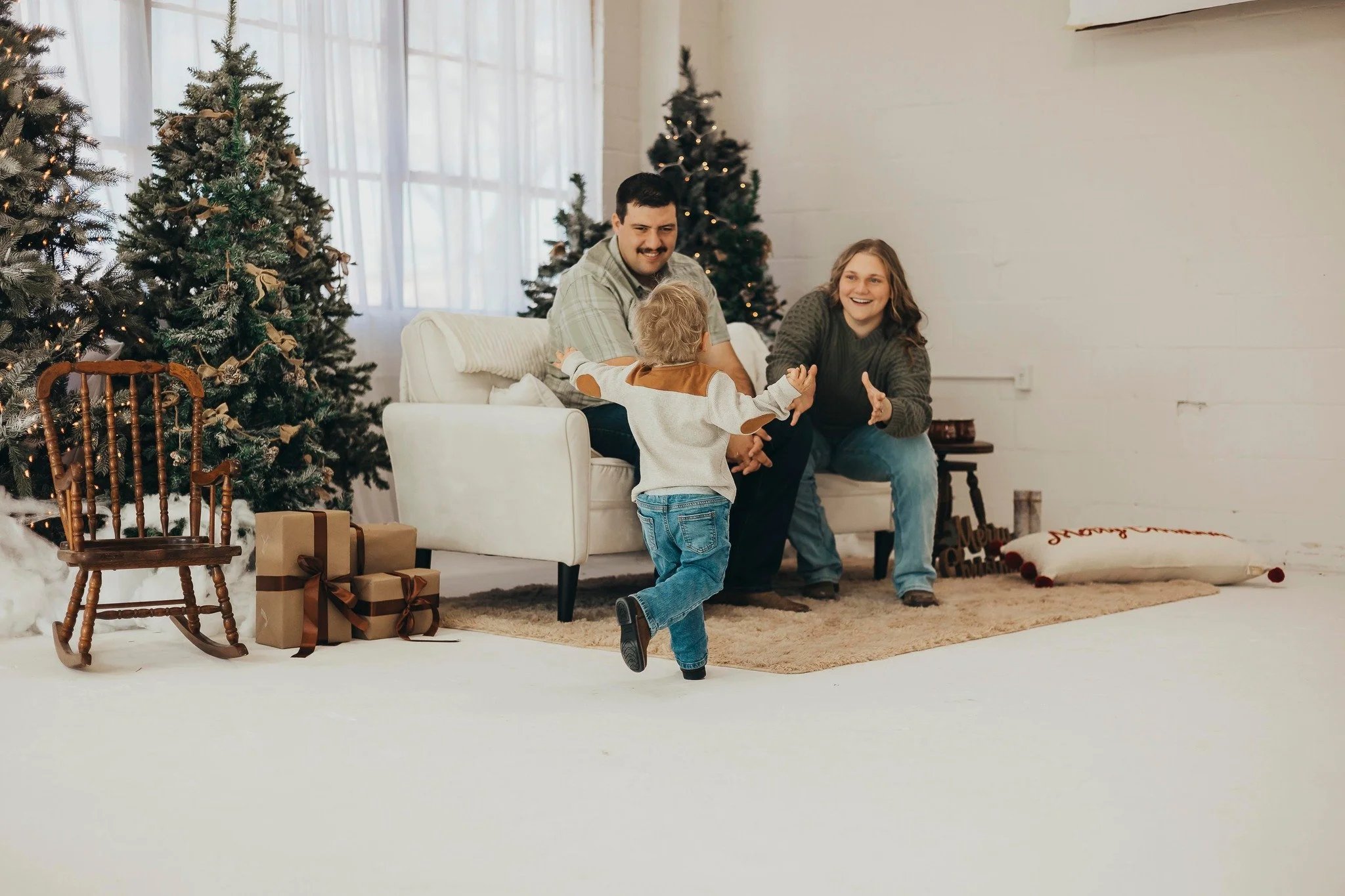 A family of three celebrating Christmas indoors. A smiling man and woman are sitting on a white sofa, while a young child runs towards them with outstretched arms. Christmas trees decorated with ornaments and lights, wrapped presents, and holiday decorations are visible in the background.