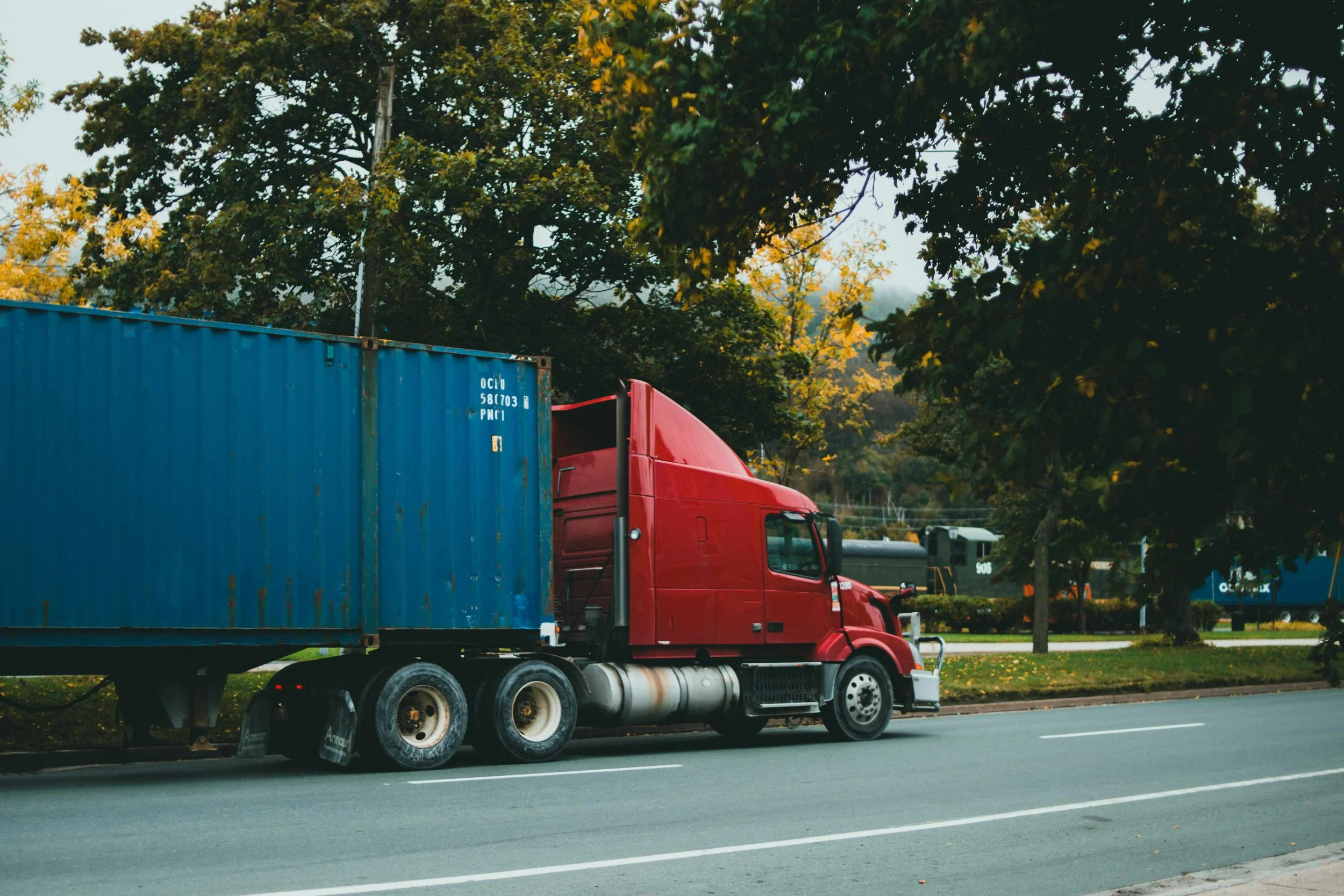 Transport truck with red cab and blue container driving on a tree lined street