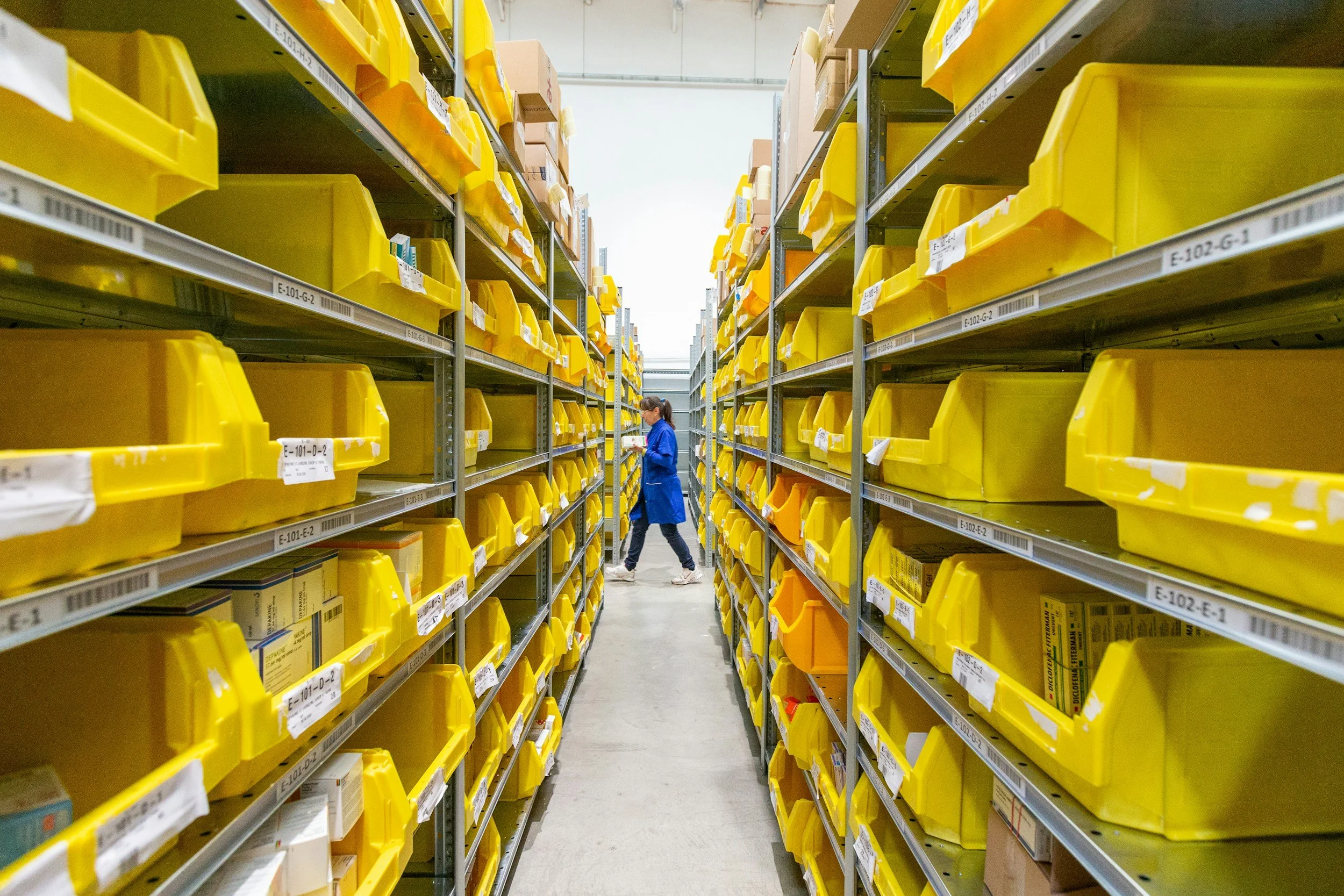 A worker in a blue uniform shopping aisle with yellow storage bins on metal shelves filled with various products.