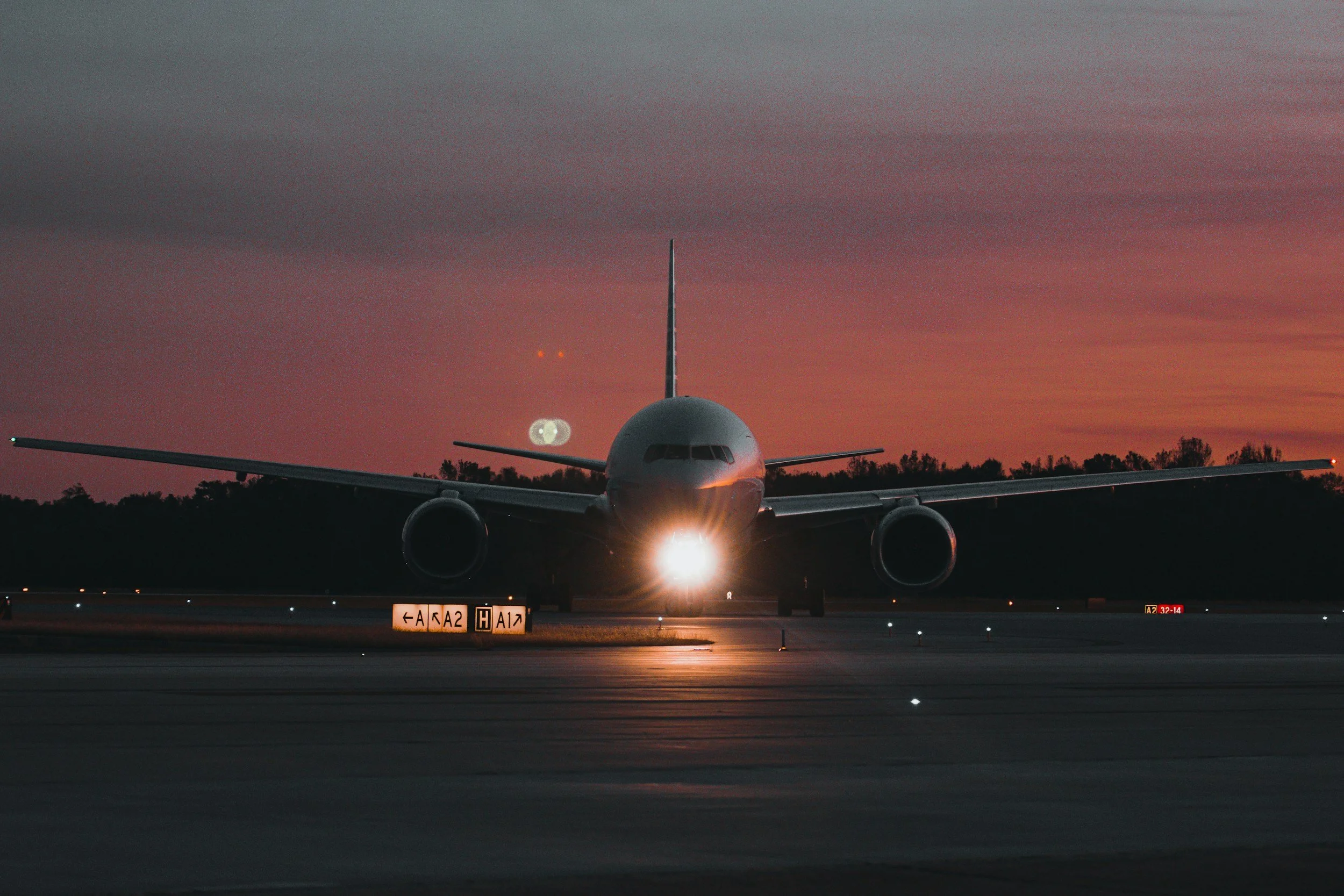 A large commercial airplane on an airport runway at sunset, facing directly toward the camera with the nose lights on, dark sky with a pinkish hue, and trees in the background.