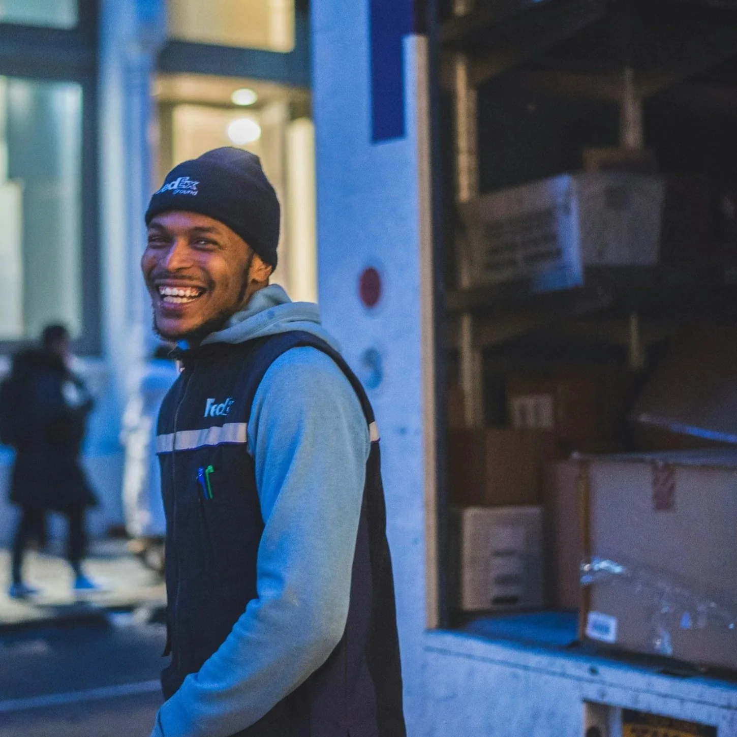 A smiling delivery man worker in a black vest and beanie, standing outdoors at dusk near a warehouse with boxes.