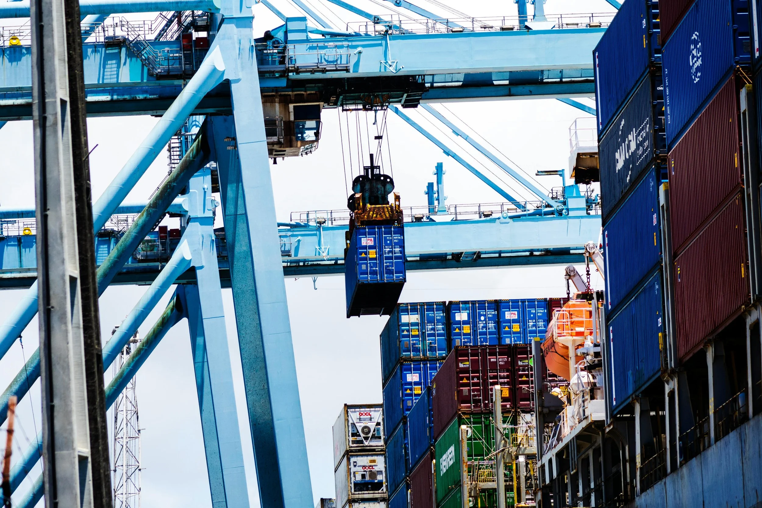 Cargo containers stacked on a shipping vessel with a large blue crane overhead loading or unloading containers.