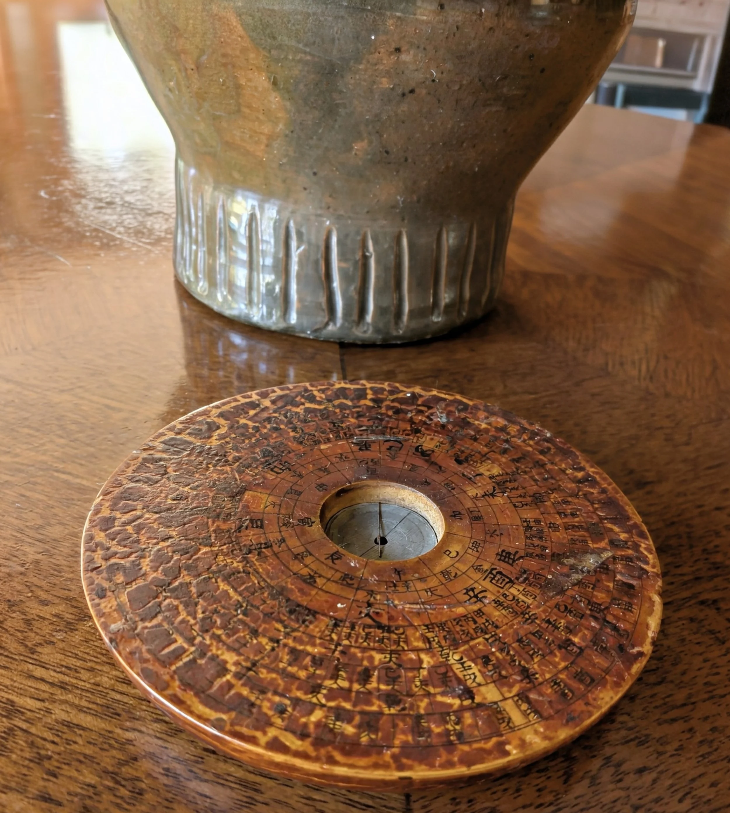 Close-up of an ancient Chinese or Asian compass and a ceramic pot on a wooden table. The compass has a weathered, orange-brown surface with Chinese characters and markings. The ceramic pot has a textured surface with lines and a slightly flared shape.