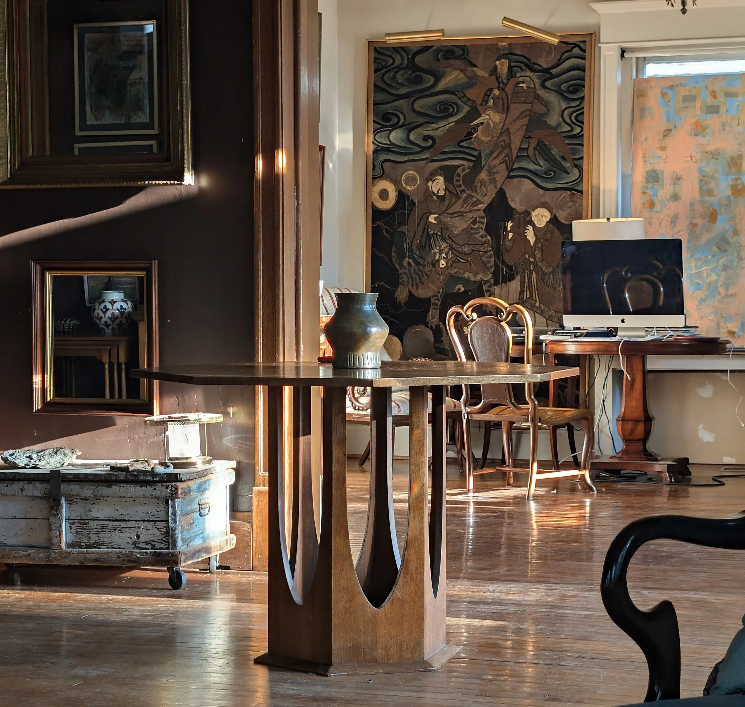 A cozy room with a wooden table displaying a ceramic vase, a vintage wooden chest on wheels, framed artwork on the walls, and a computer on a wooden table near a window.