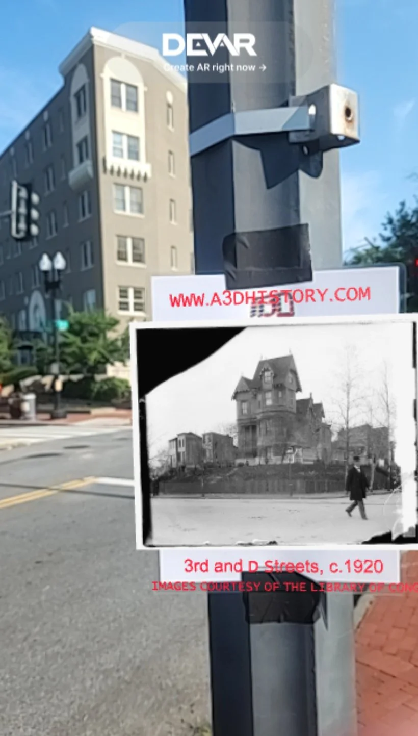 A street corner with a historical black-and-white photograph of old houses posted on a metal pole, with modern buildings visible in the background and an online website URL at the top.