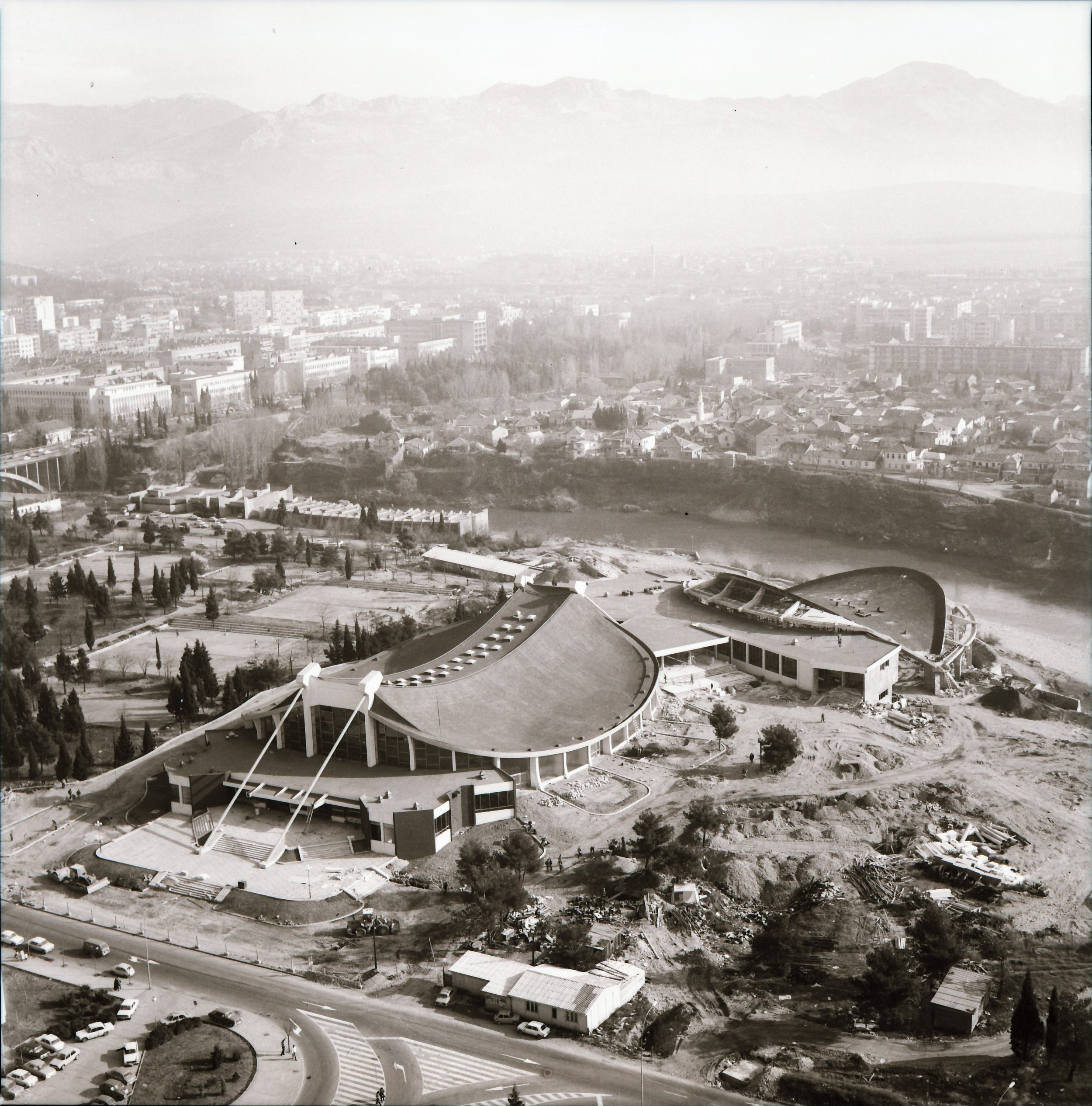 Black and white aerial photo of the construction site of the Utah Olympic Oval, with a large building featuring a curved roof, construction vehicles, and nearby roads and landscape.