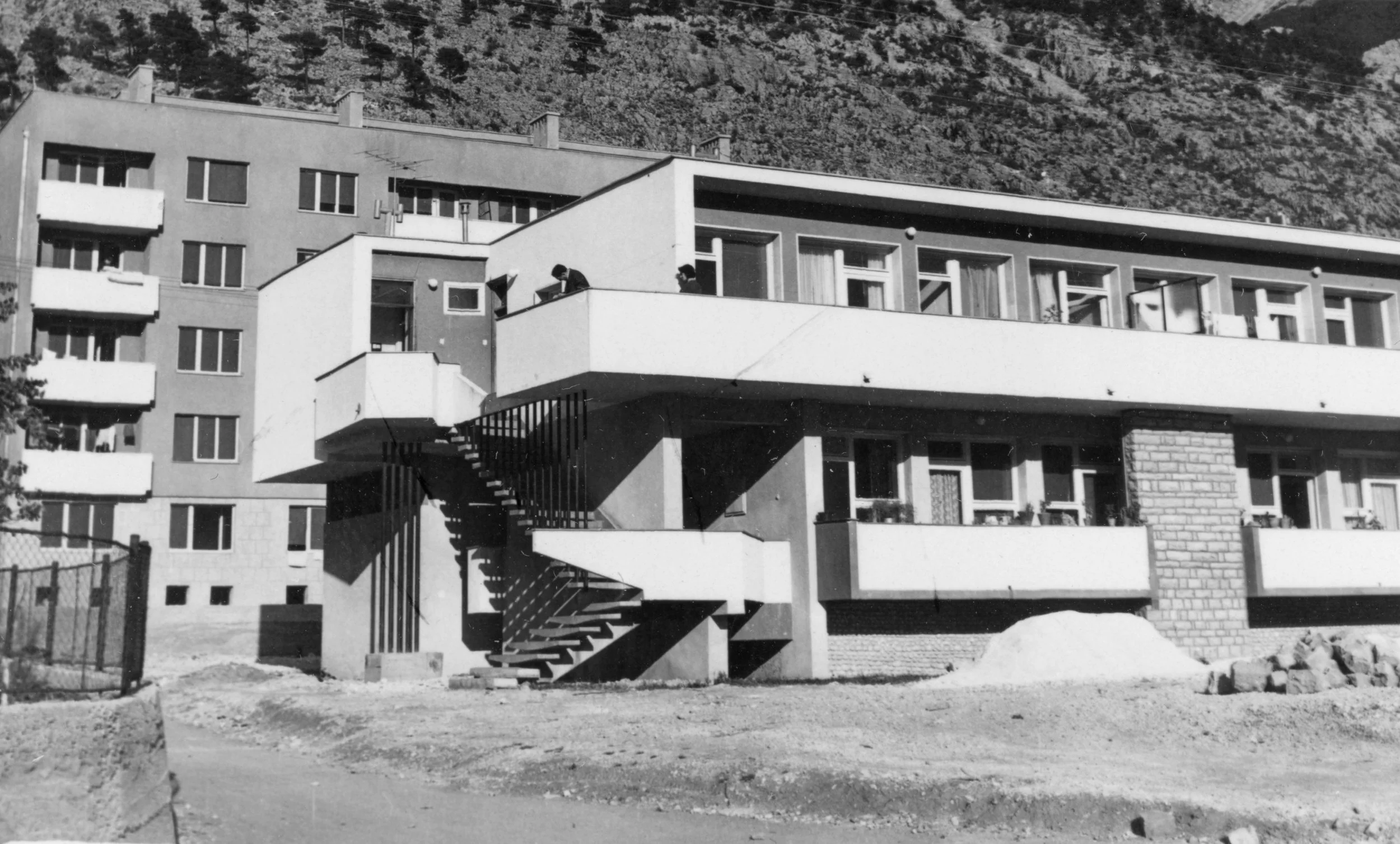 Black and white photo of a modern multi-story residential building with balconies and an external staircase, set against a hillside with trees.
