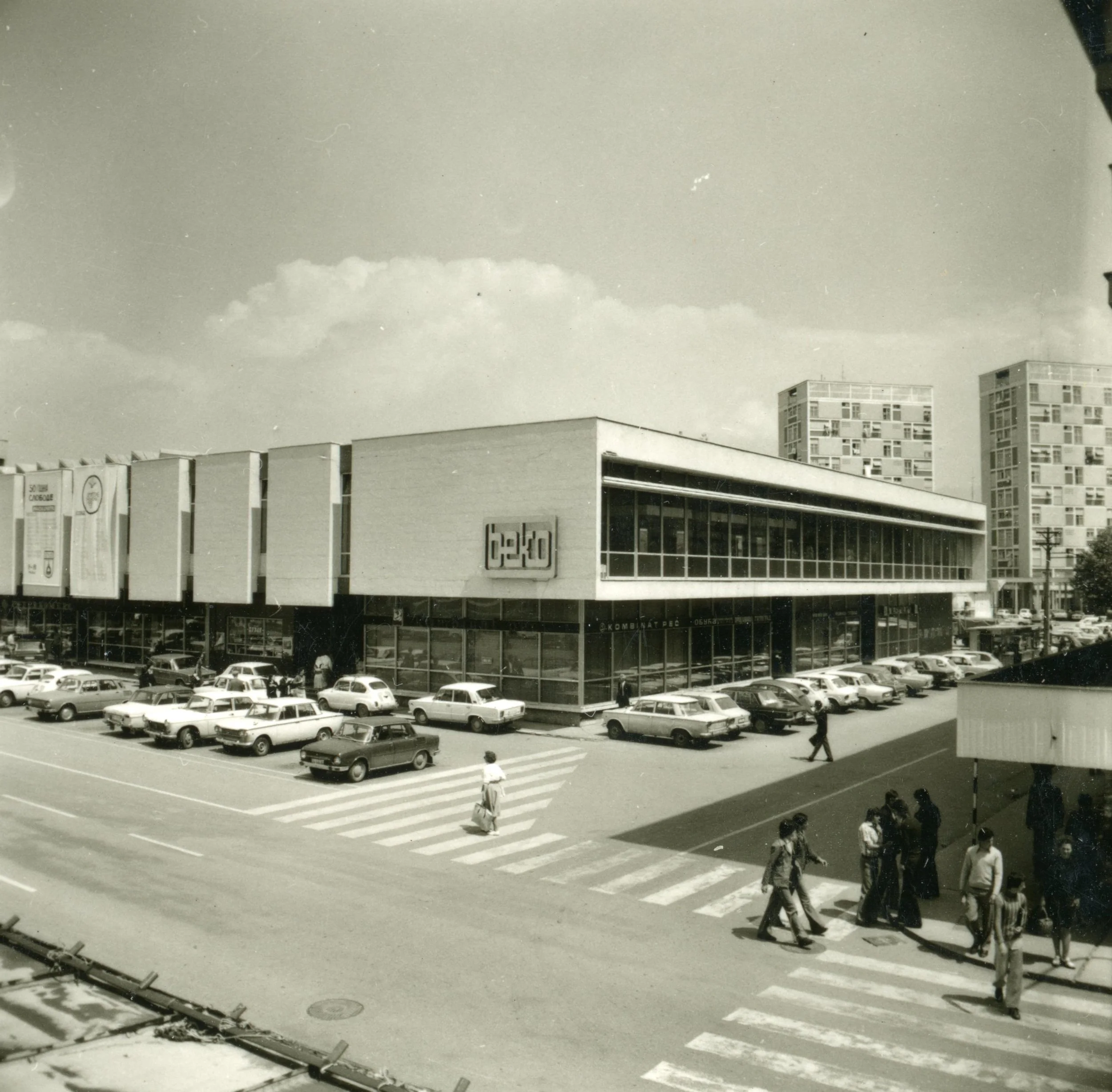 Black and white photo of a shopping center with a parking lot filled with cars, people walking on the crosswalk, and pedestrians near the entrance. The building has large glass windows and a sign that says 'beko'.