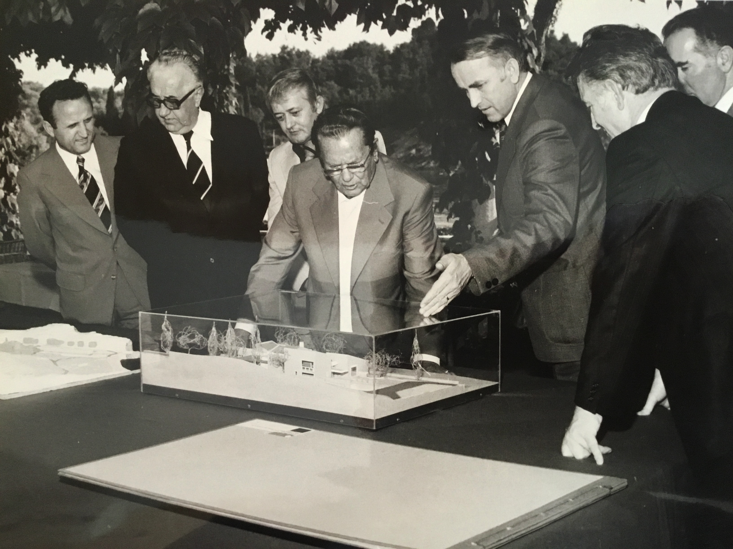 A group of men in suits and ties gathered around a table, examining a detailed architectural model encased in glass. They appear to be engaged in a discussion about the model, which is set outside under a tree.