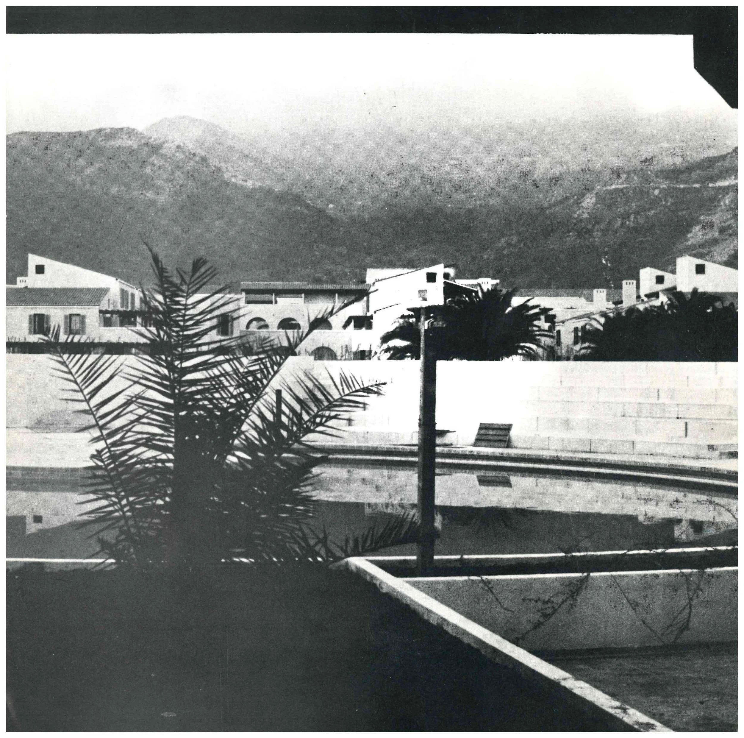 A black-and-white photo of a courtyard with a rectangular pool in the foreground, a bench, and a palm tree beside the pool. In the background, there are white buildings with arched windows and balconies, surrounded by palm trees. Mountains are visibl