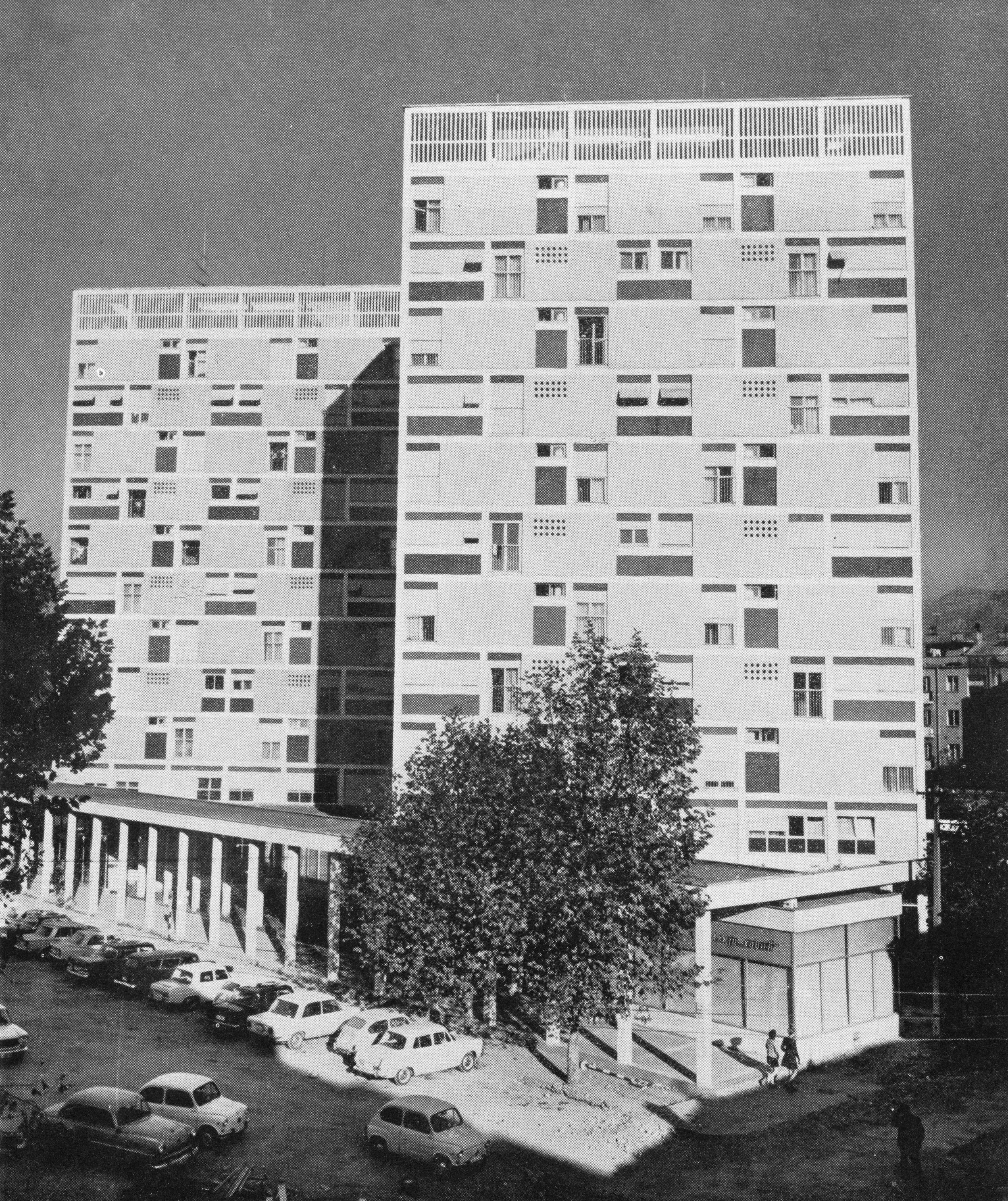Black and white photo of a modern residential high-rise building with a parking lot and trees in the foreground.
