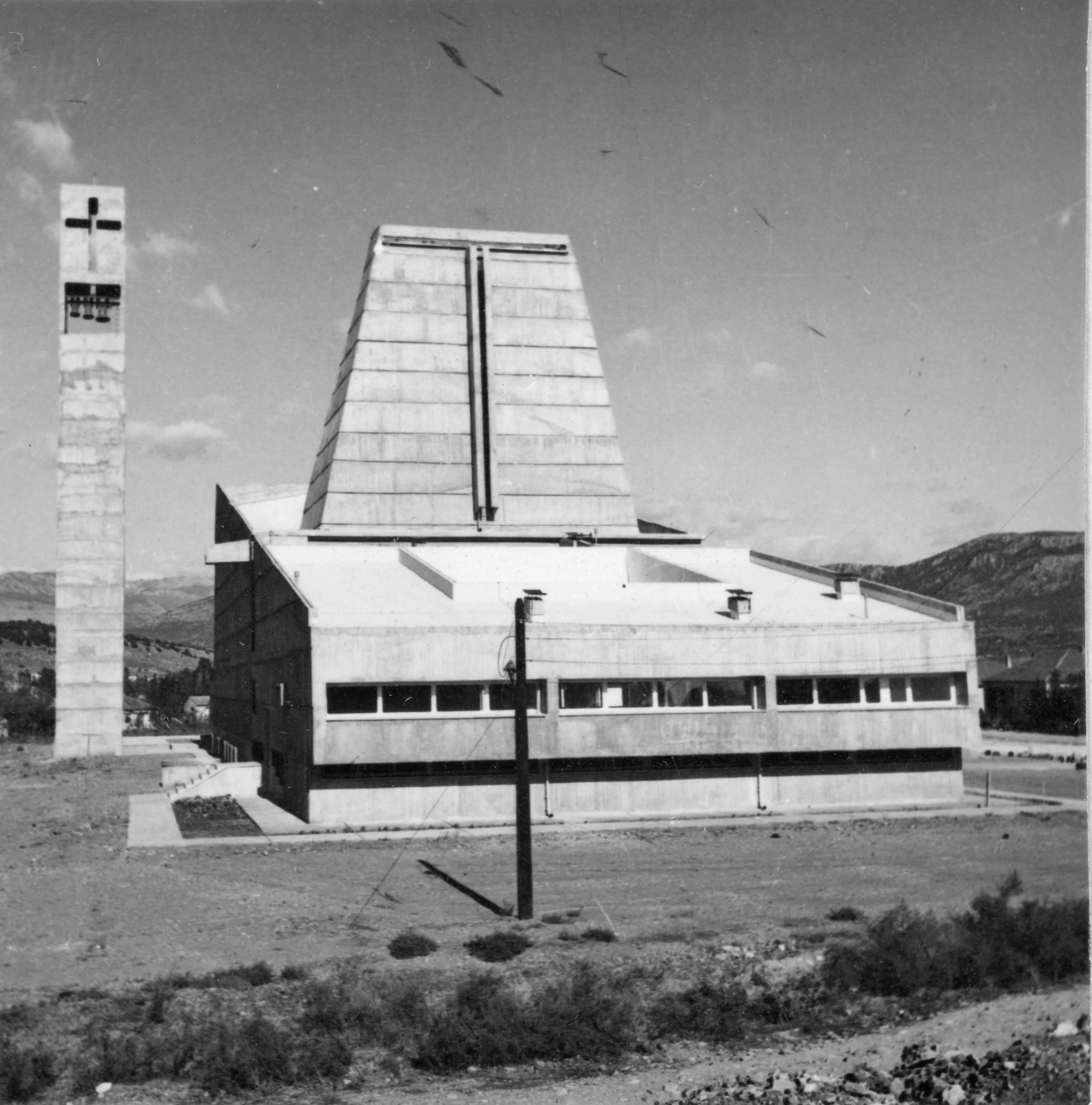 A black-and-white photo of a modernist church with a tall, pointed steeple and horizontal windows, set in a rural landscape with mountains in the background.