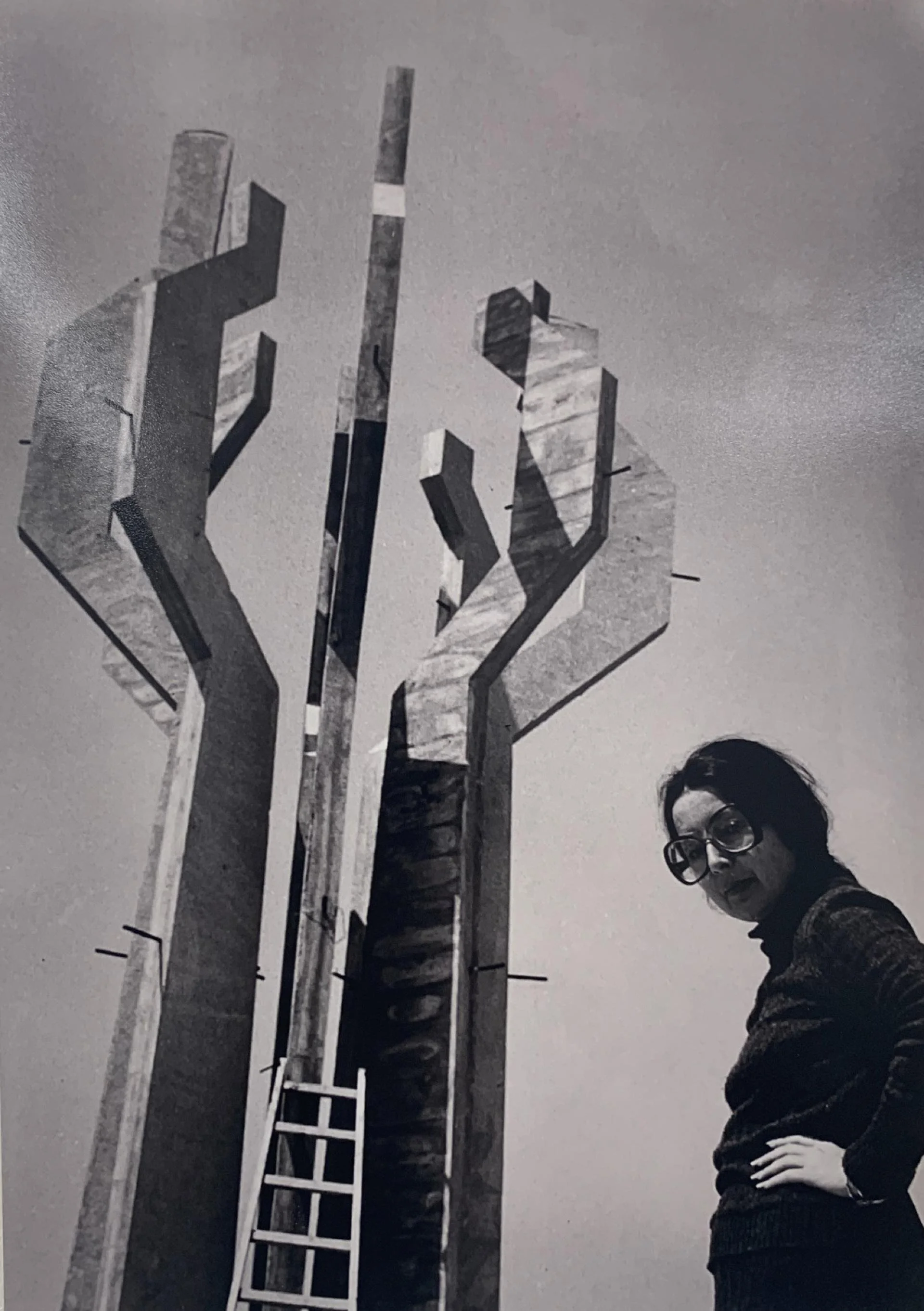 A black and white photograph of a woman with glasses and dark hair standing beside a towering modern sculpture resembling hands making the 'peace' or 'victory' sign, set against a cloudy sky.