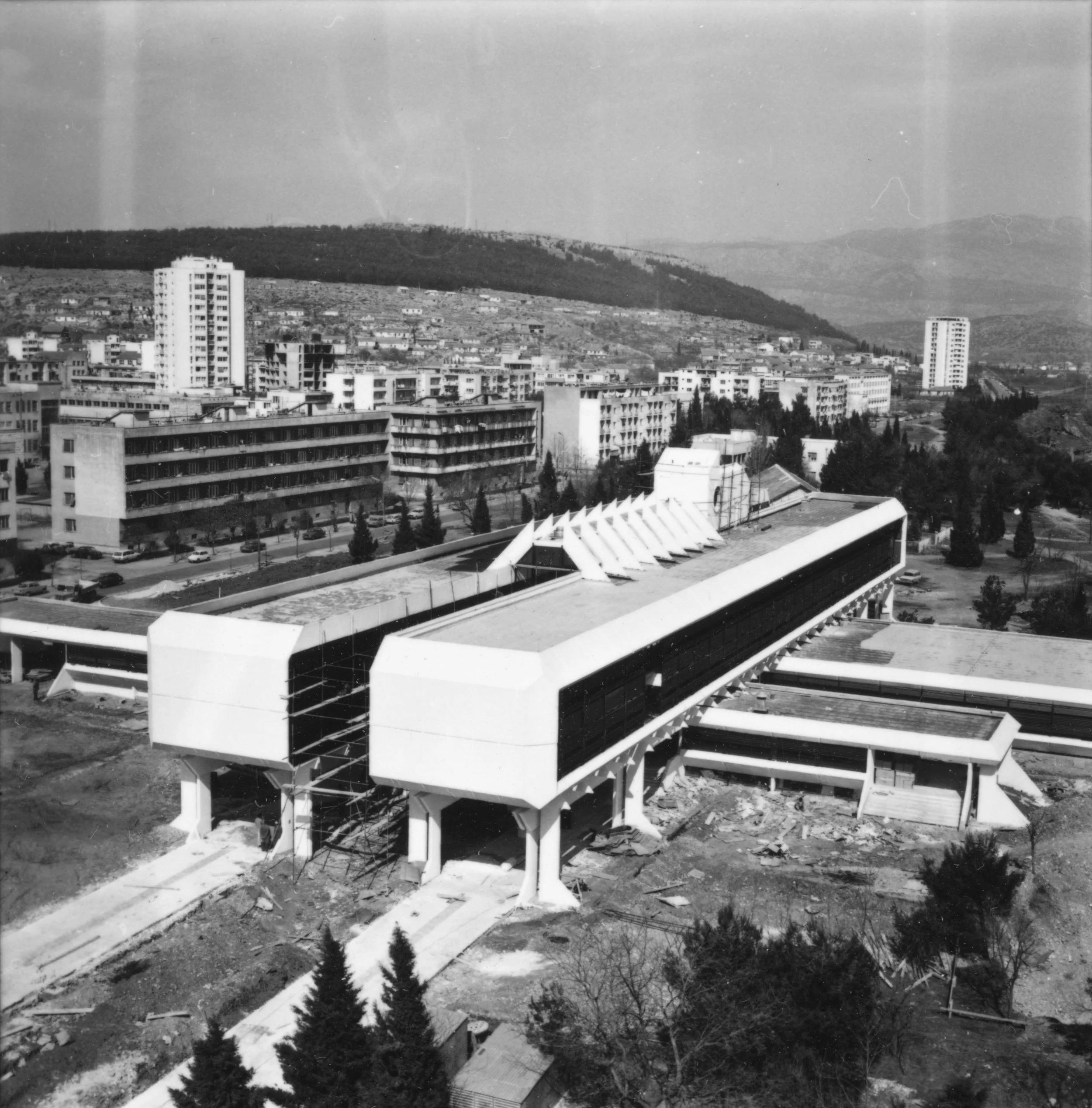 Black and white photo of a futuristic building under construction with a long, black exterior and white structural elements, situated in an urban area with scattered trees and multiple apartment buildings in the background, hills in the distance.