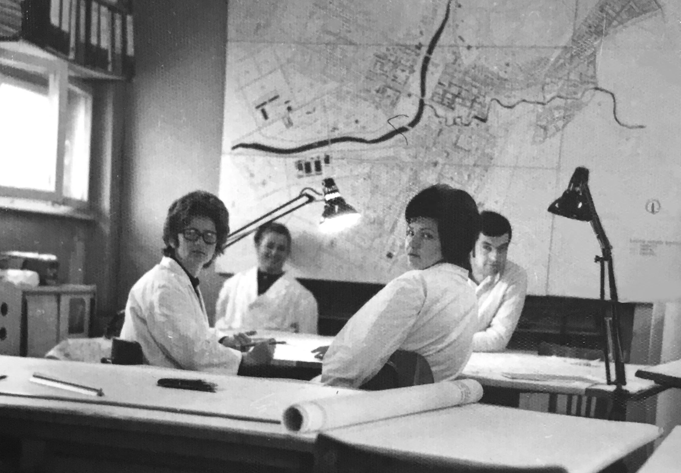 Black and white photo of four people in a meeting or planning session, sitting around a desk with documents and writing tools. A large map is on the wall behind them, and two desk lamps are illuminating the workspace.
