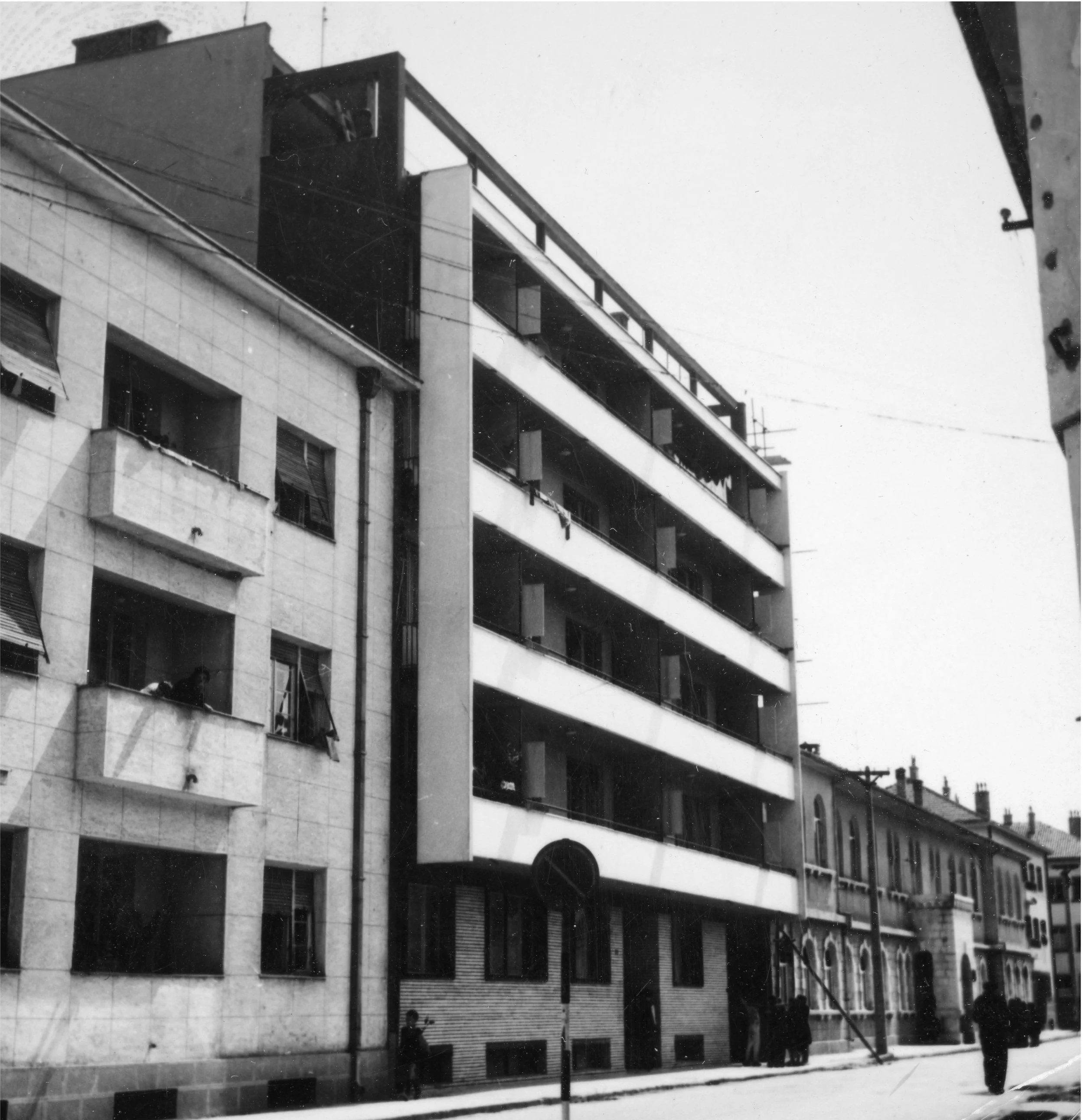 Black and white photo of a multi-story apartment building on a city street with a sidewalk and pedestrians.