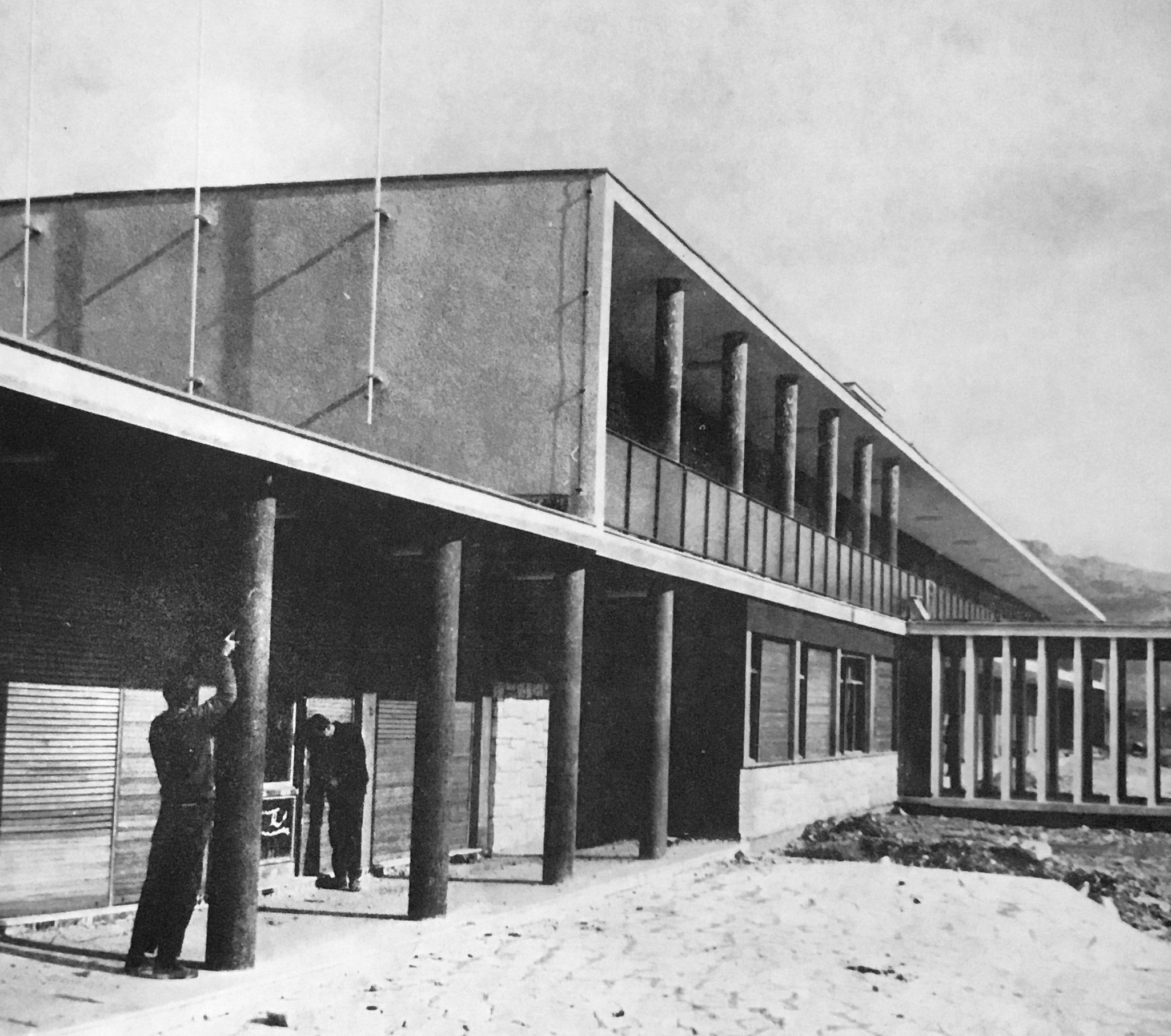 Black and white photo of a modern building under construction with tall support columns, scaffolding, and construction workers.