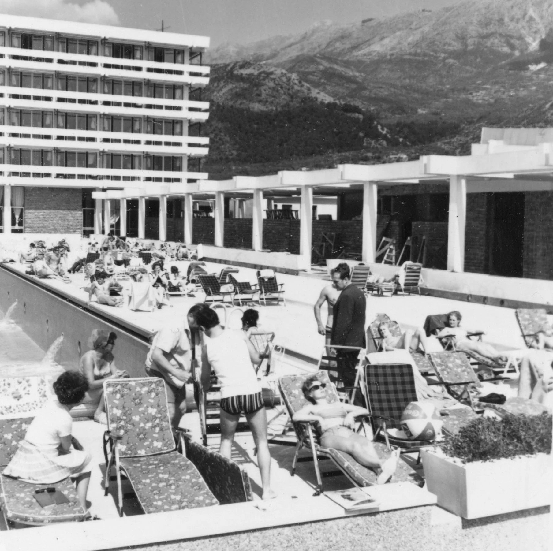 Black and white photograph of a hotel poolside scene with multiple people lounging on chairs, some swimming, and others talking, with a mountainous background.