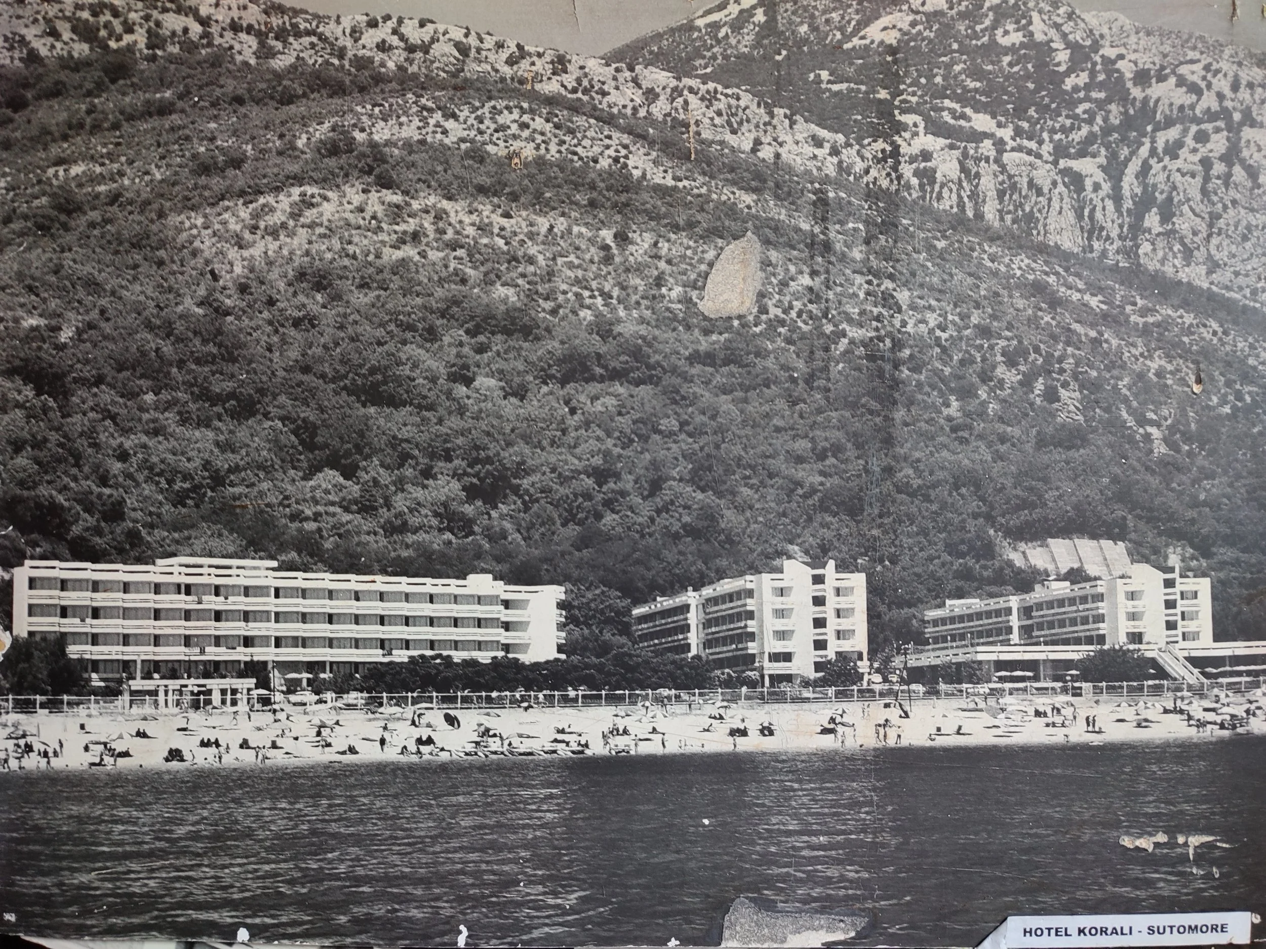 Black and white photo of a beach with umbrellas and people, three large hotel buildings in the background, with a mountain covered in trees behind the hotels.