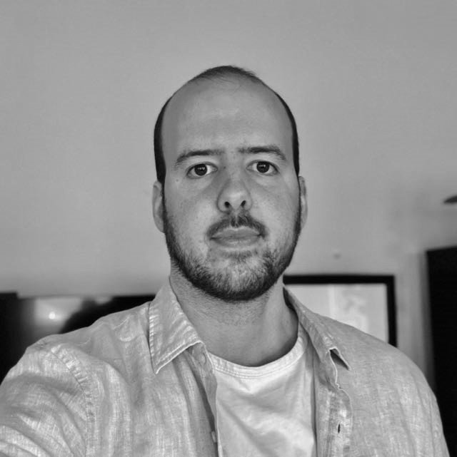 Black and white photo of a man with a beard and short hair, wearing a light-colored shirt and white t-shirt, standing indoors with a framed picture in the background.