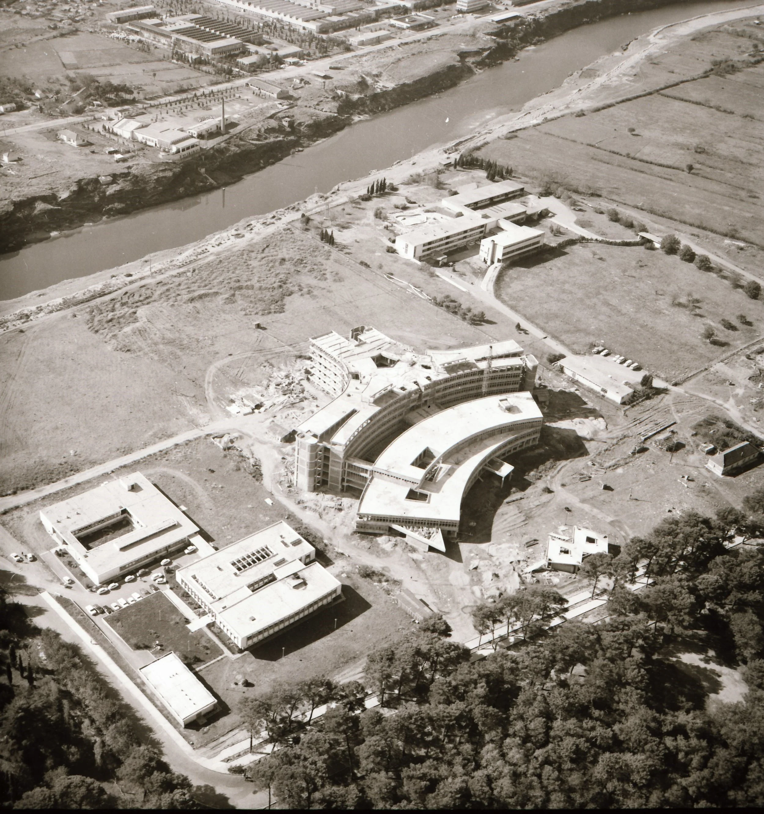 Aerial view of a construction site with a large, curved building under development, surrounded by scattered smaller structures and greenery, near a river.