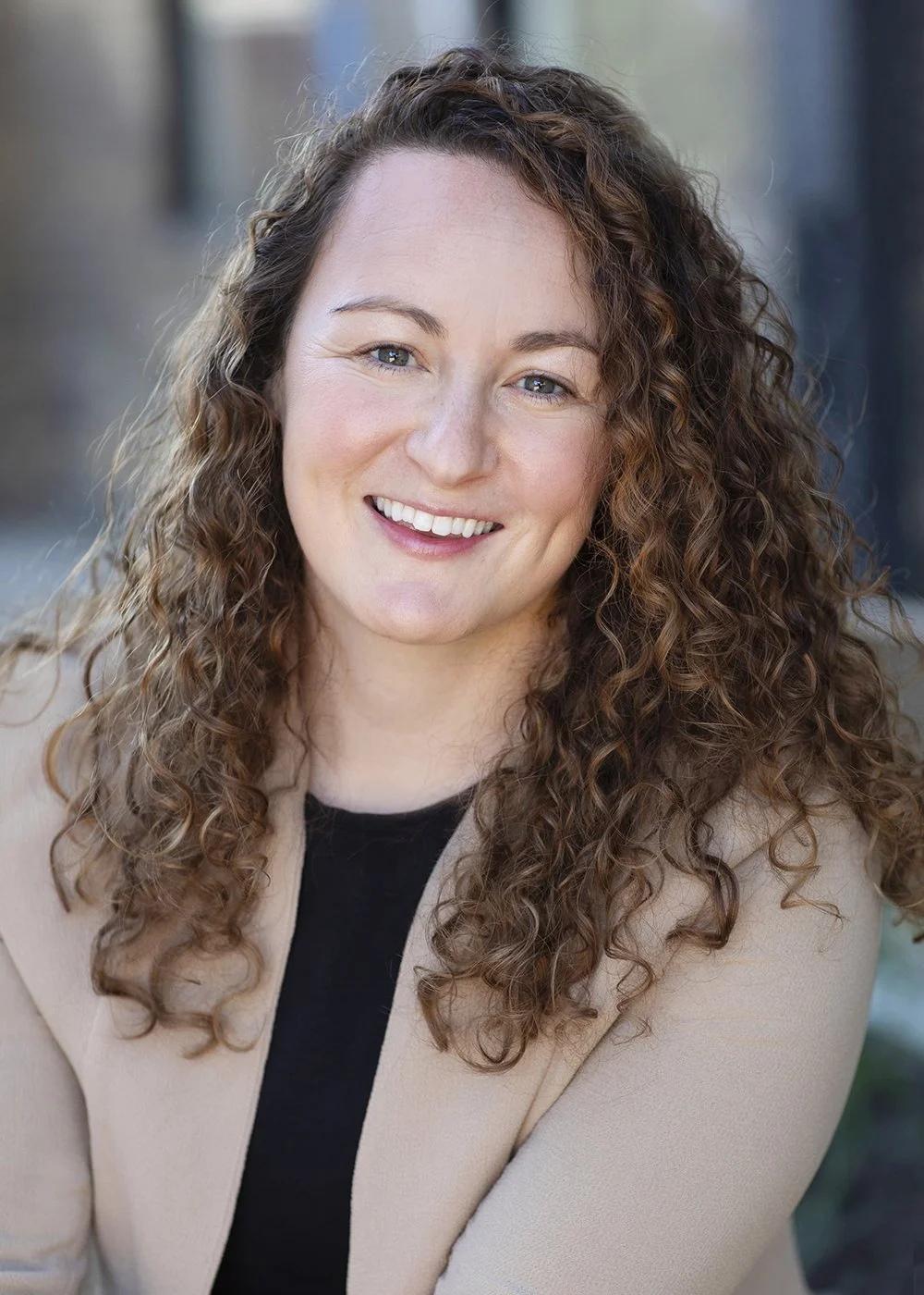 A woman with long, curly brown hair smiles outdoors with a blurred urban background.