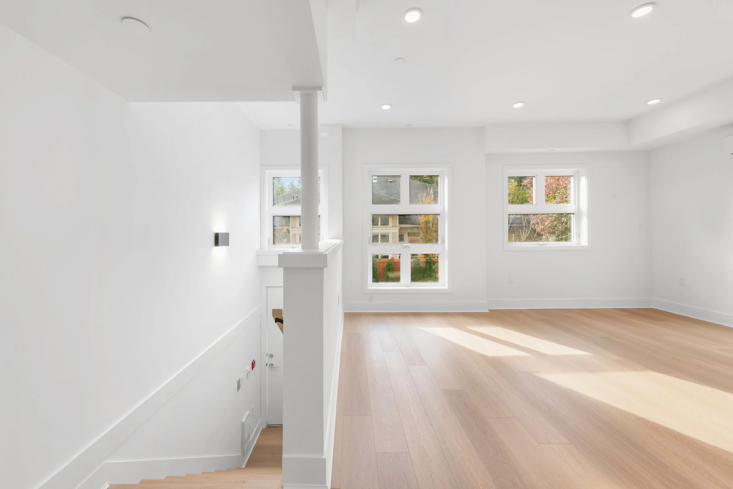 Bright empty room with large windows, white walls, and light wooden floors, with stairs visible to the left.