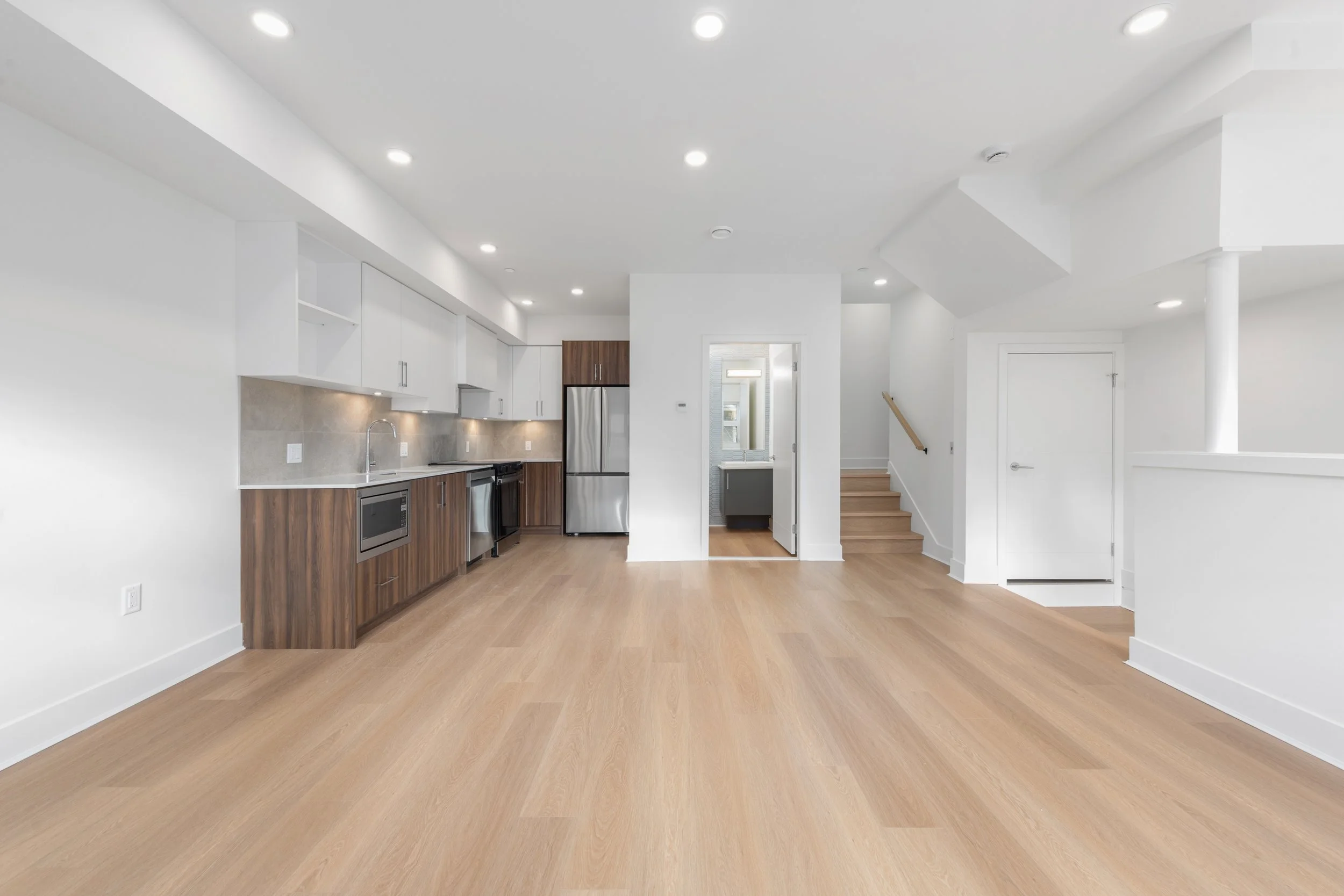 Empty modern kitchen with white upper cabinets, brown lower cabinets, stainless steel appliances, and light wood flooring.