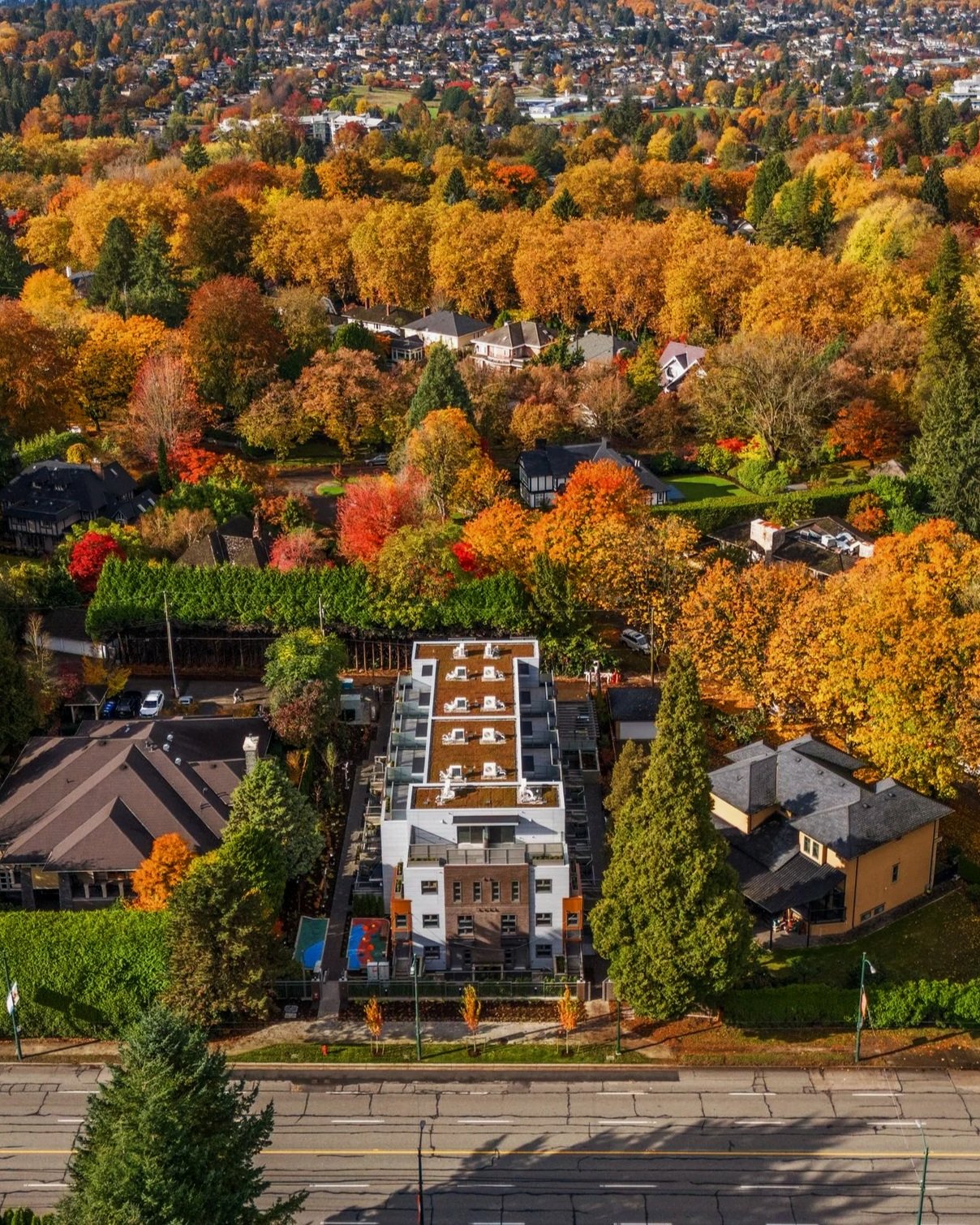An aerial view of a neighborhood during fall, showing colorful autumn trees and a modern multi-story residential building in the foreground.