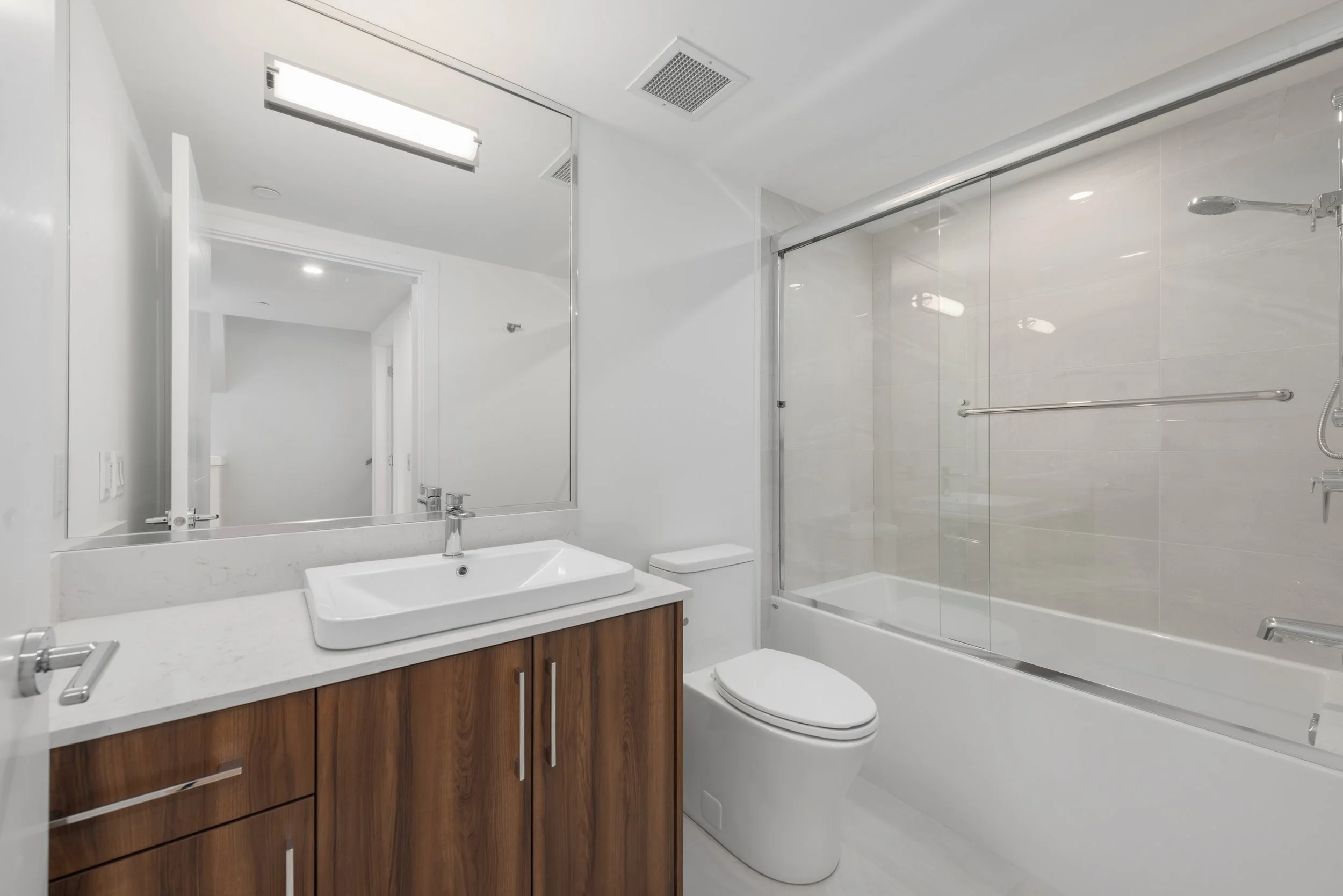 Modern bathroom with white walls, wooden vanity with a white sink, large mirror, and a glass-enclosed shower with a bathtub.