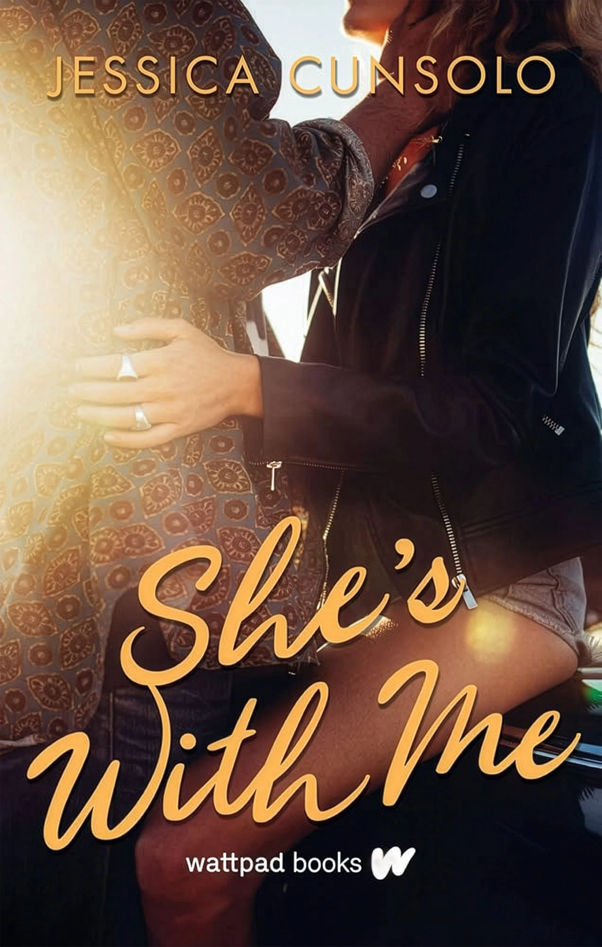 Book cover for 'She's With Me' by Jessica Cunsolo published by Wattpad Books. The cover features two women embracing each other, with one woman in a patterned long-sleeve shirt and the other in a black jacket. The background shows a sunset or sunrise.