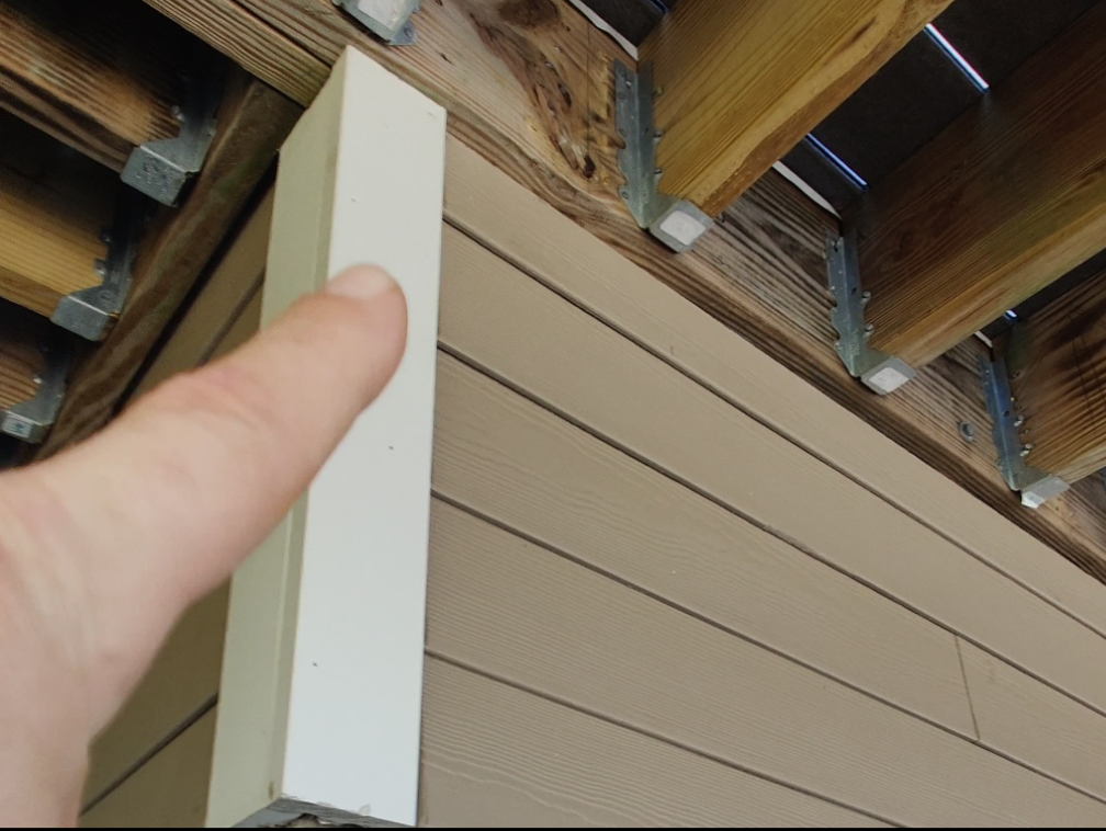 A person's finger touching a white wooden trim board on a house exterior, with siding below and a wooden deck above.