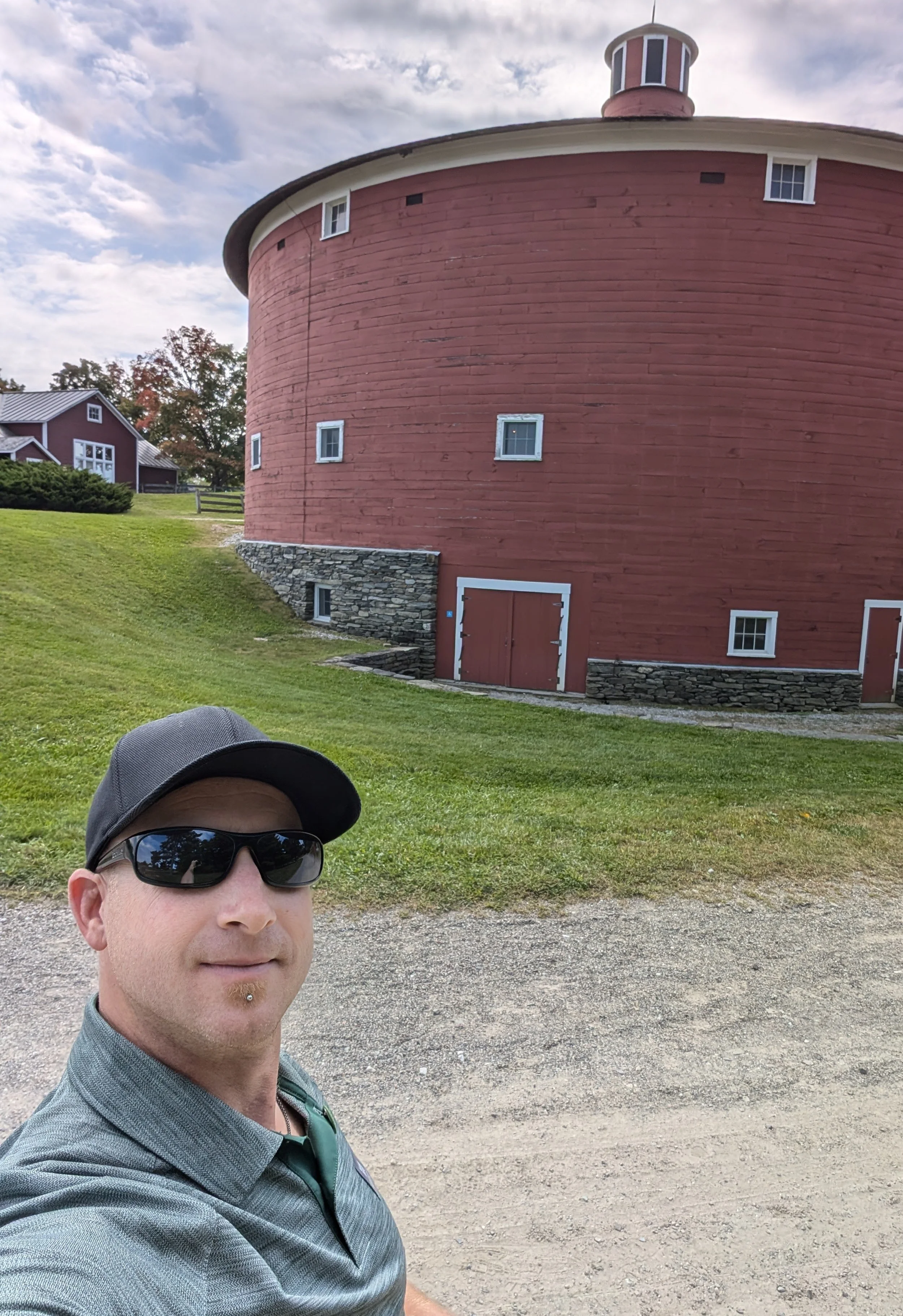 A man wearing sunglasses, a cap, and a gray shirt taking a selfie in front of a round red barn with small windows and stone foundation, under a partly cloudy sky.