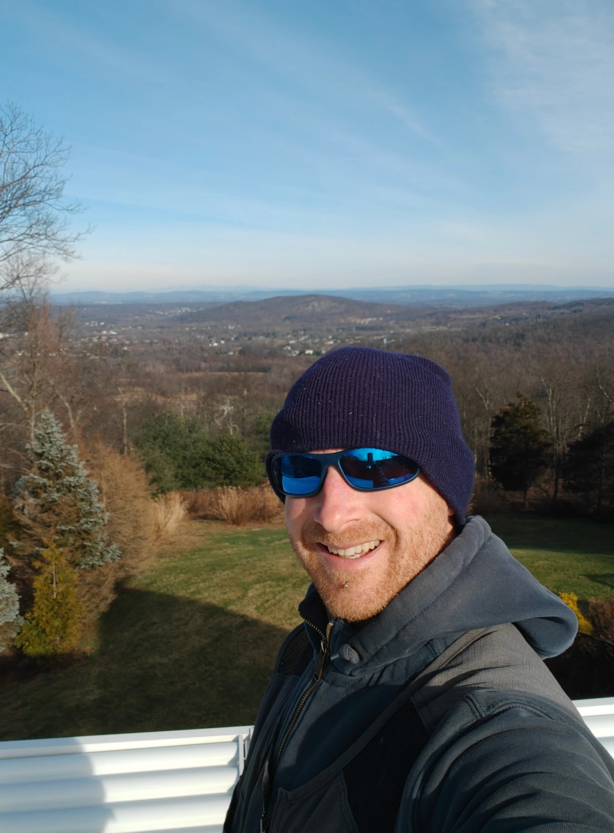 A man taking a selfie outdoors on a clear day, wearing sunglasses, a navy beanie, and a gray jacket. The background features a scenic landscape with trees, rolling hills, and a distant view of the horizon.