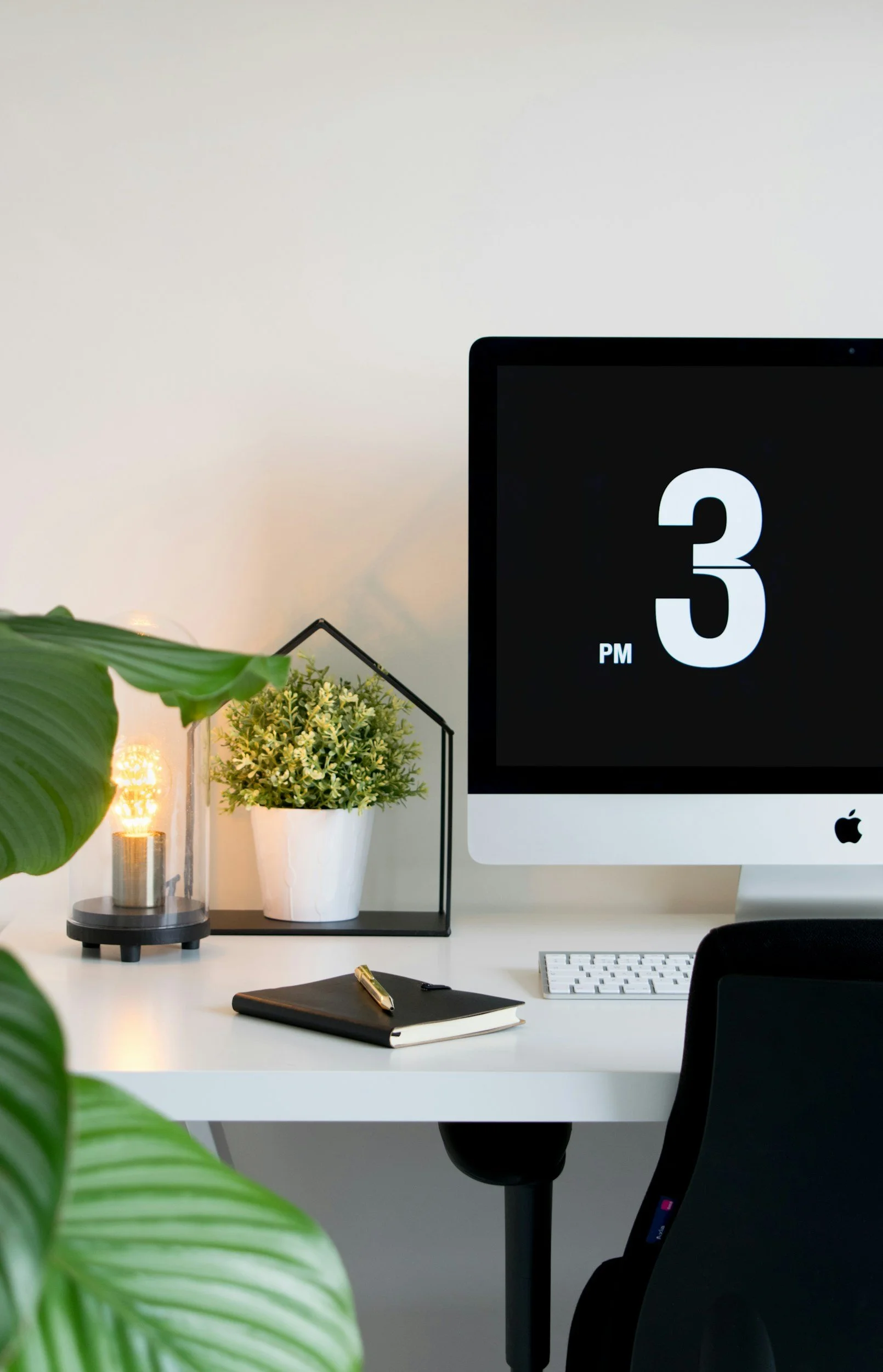 Modern office workspace with a white desk, greenery, and a computer monitor displaying the time 3 PM, surrounded by a black chair, a notebook with a pen, and decorative plant arrangements
