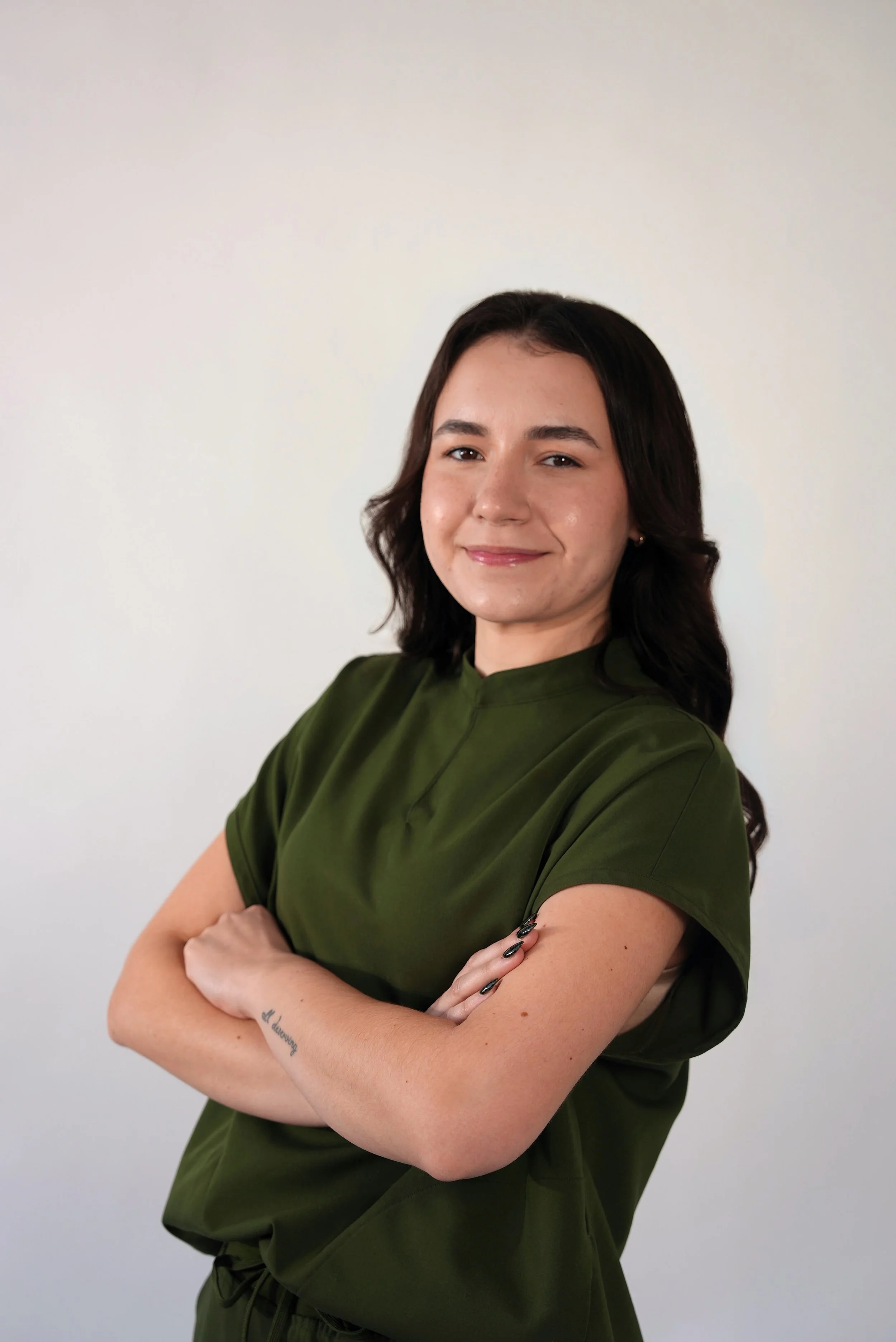 A woman with dark brown hair and fair skin standing with her arms crossed, smiling slightly against a plain white wall background, wearing a dark green scrub top.